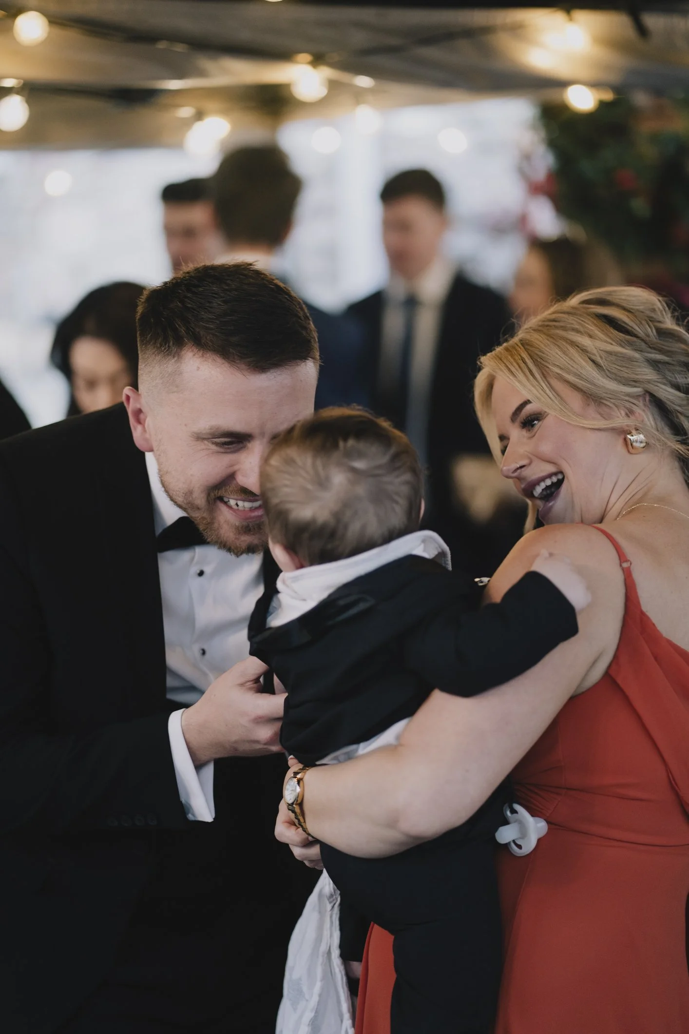 A man in a tuxedo, a woman in a red dress, and a young child sharing a happy moment at a wedding at Radnor Rooms