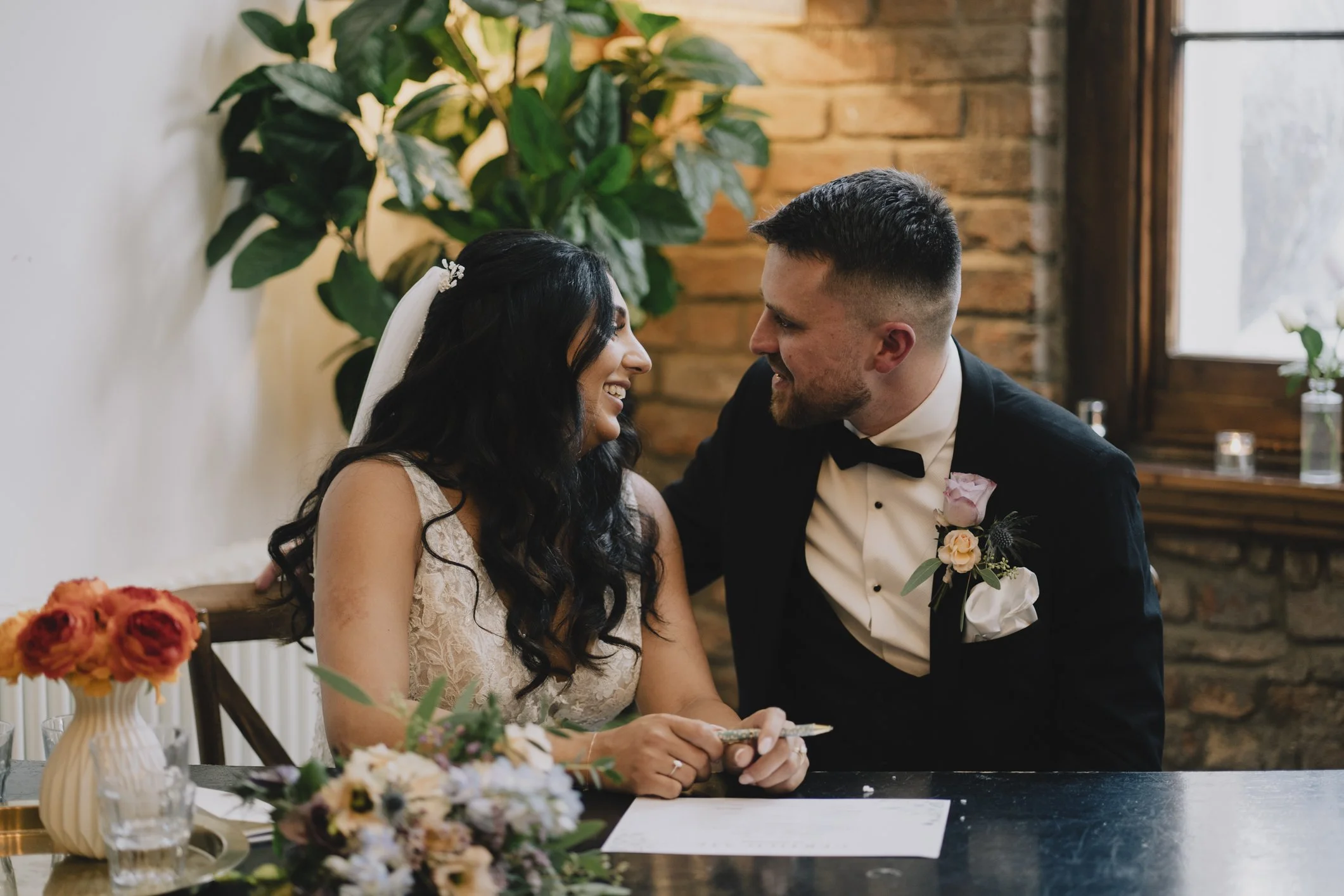 A newlywed couple smiling and gazing at each other during their wedding ceremony, sitting at a table with flowers, in a room with brick wall and large window in Bristol