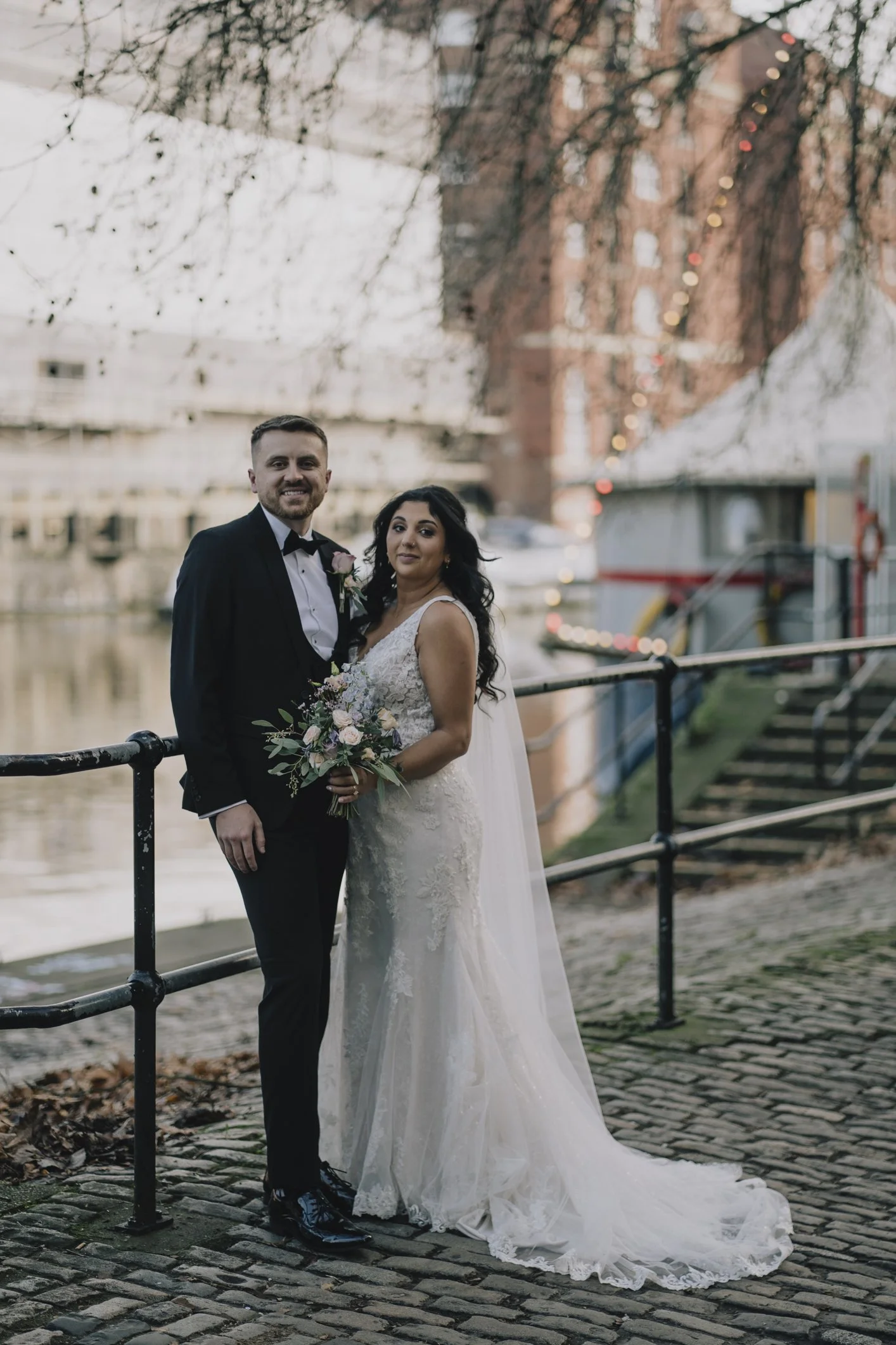 Wedding couple standing by river on cobblestone path with bridge, buildings, and trees in the background in Bristol