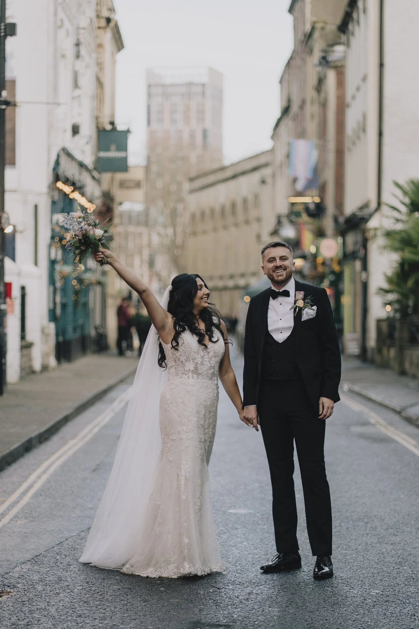 A newlywed couple is walking hand in hand on a city street. The bride, dressed in a white lace wedding gown, is smiling and holding a bouquet in the air. The groom, in a black tuxedo, is smiling. They are surrounded by buildings and a few pedestrians in the background