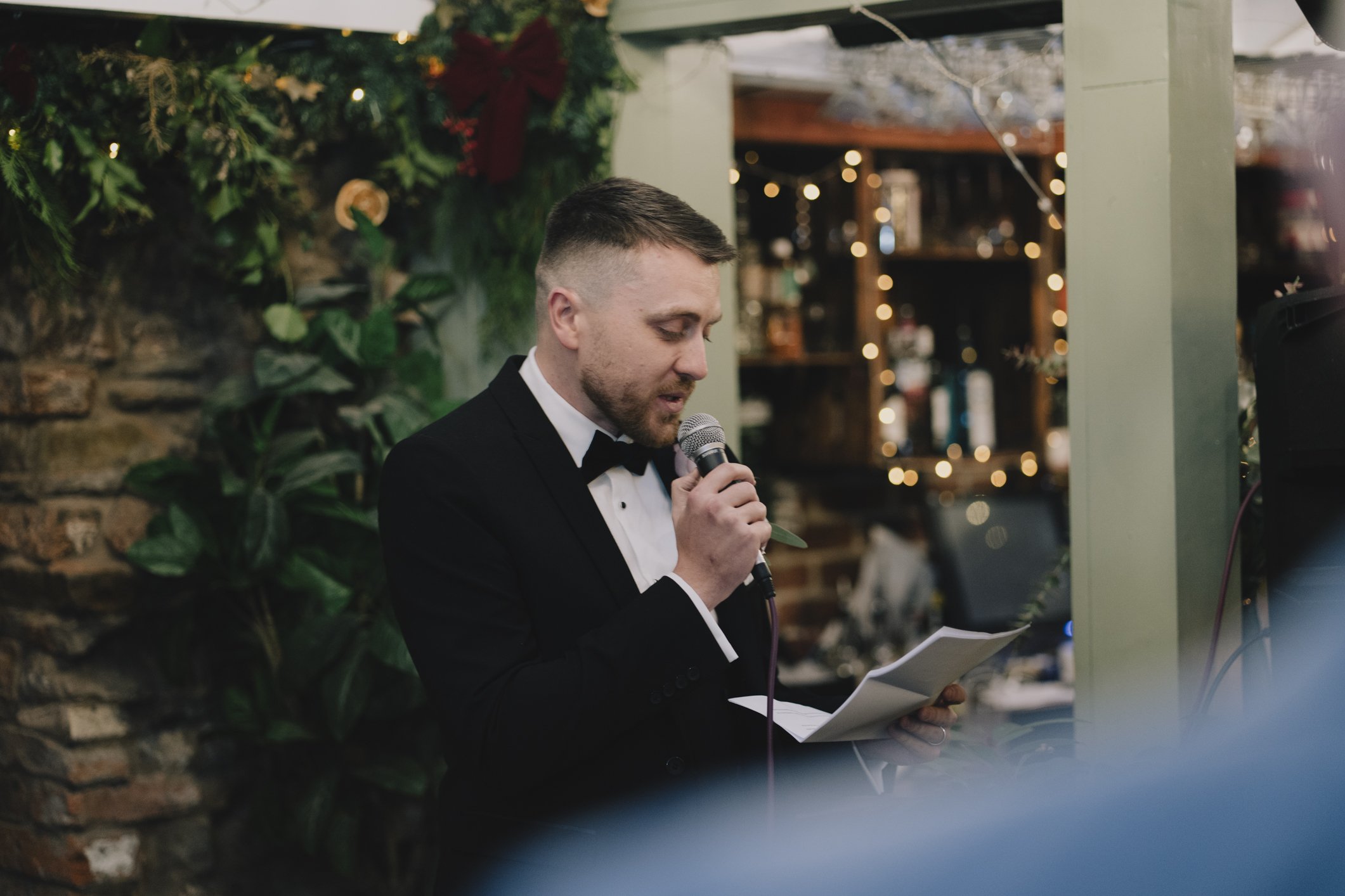 Man in a tuxedo speaking into a microphone while reading from a piece of paper at a decorated Radnor Rooms
