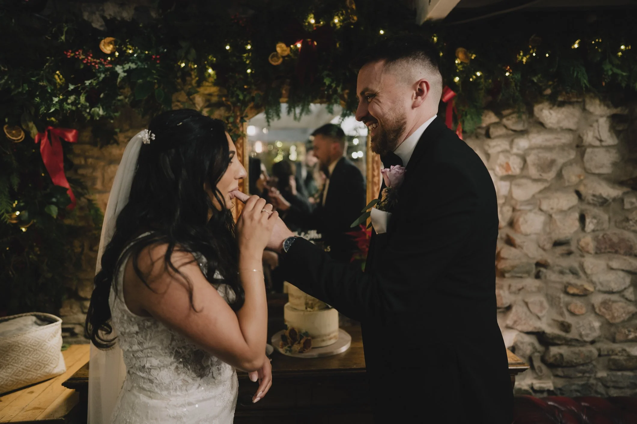 A wedding couple standing close together indoors, with the groom smiling and holding the bride's hand. The bride is wearing a lace wedding dress and veil, and the groom is dressed in a black tuxedo with a boutonniere. There is a decorated stone wall and foliage with fairy lights and red ribbons in the background.