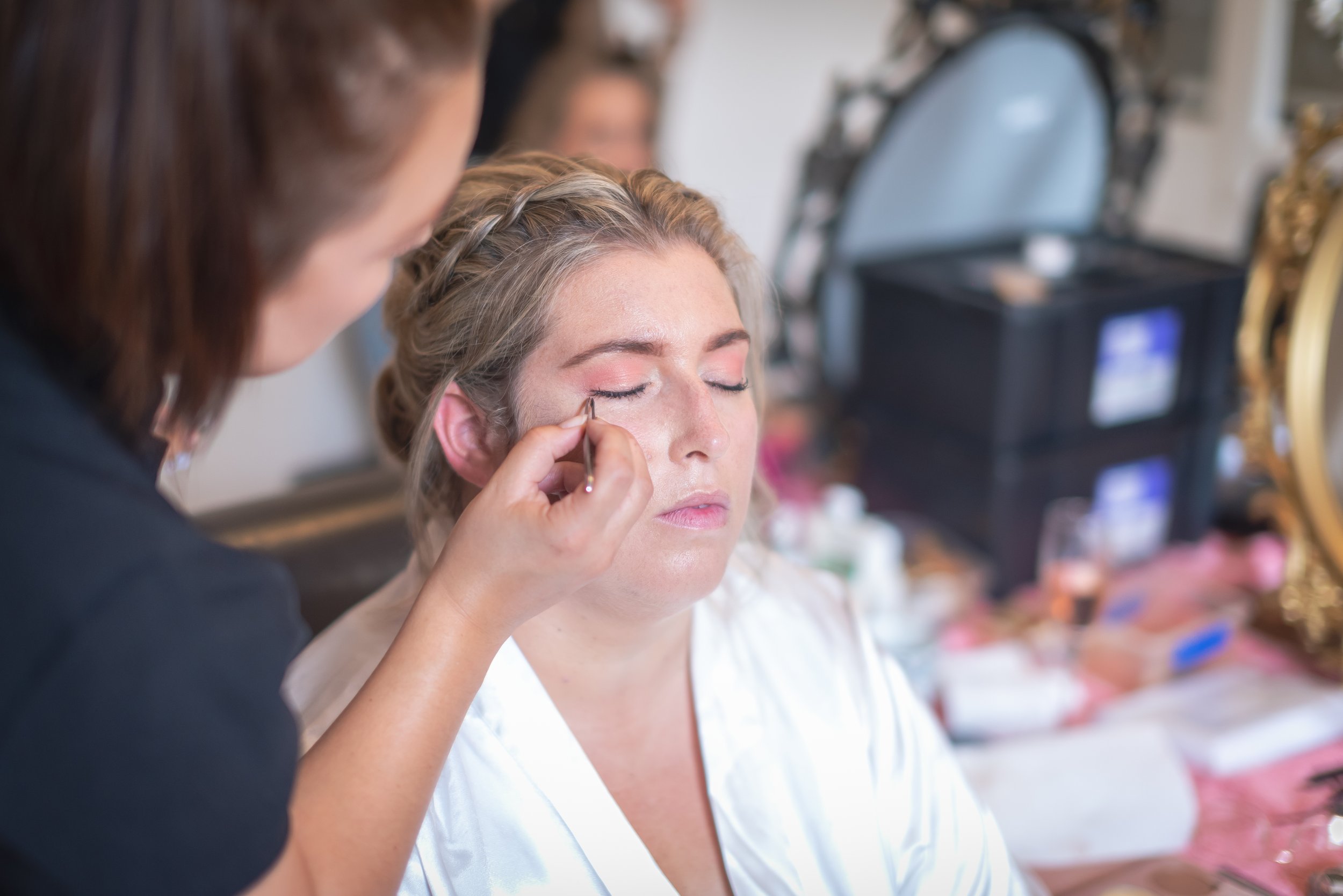 A woman with light skin and curly blonde hair has her eyes closed as a makeup artist applies eyeliner to her eyelids.