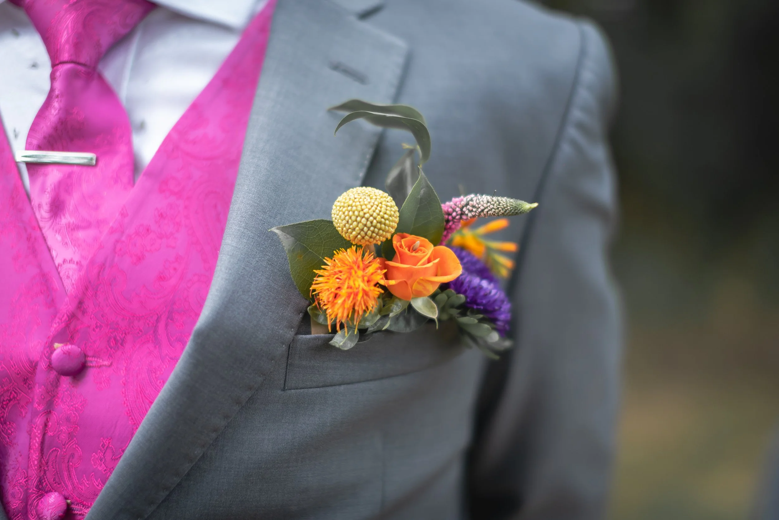 Close-up of a gray suit jacket with a vibrant flower boutonniere, including yellow, orange, purple, and white flowers and green leaves, worn over a pink patterned vest and white shirt.