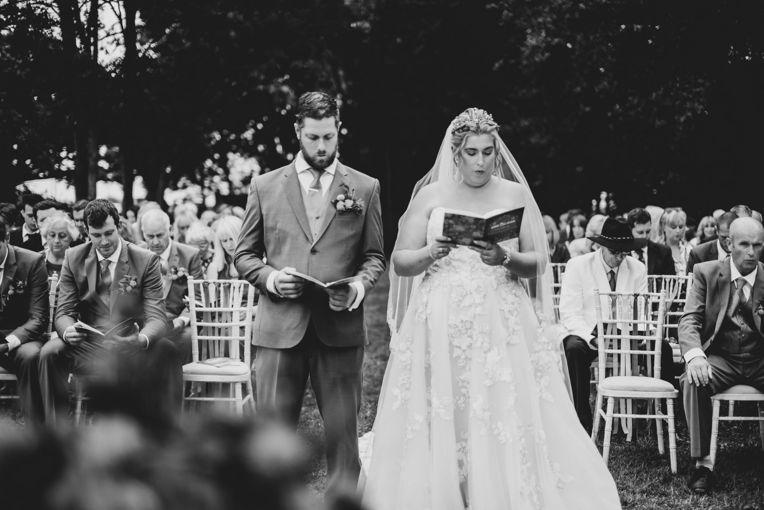 A black and white photo captures a bride and groom standing during their outdoor wedding ceremony. The bride is wearing a strapless wedding gown with floral details and a veil, reading from a book. The groom is in a suit, also holding a book. Guests are seated behind them, paying attention to the ceremony, with some dressed in formal attire and one woman wearing a large hat.