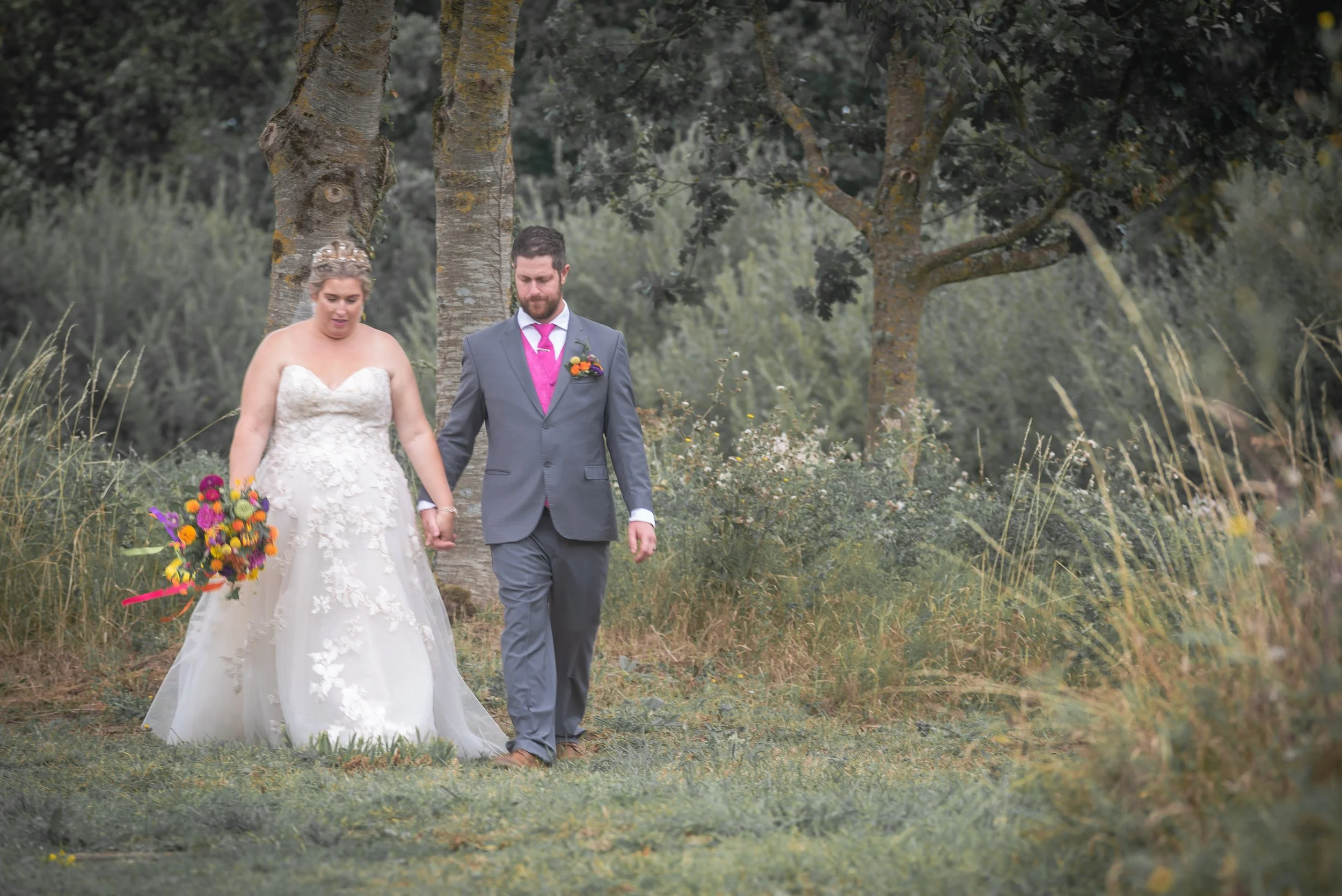 A bride and groom walking hand in hand outdoors in a natural setting with trees and grass, the bride holding a colorful bouquet of flowers at Thorney Lakkes during wedding reception
