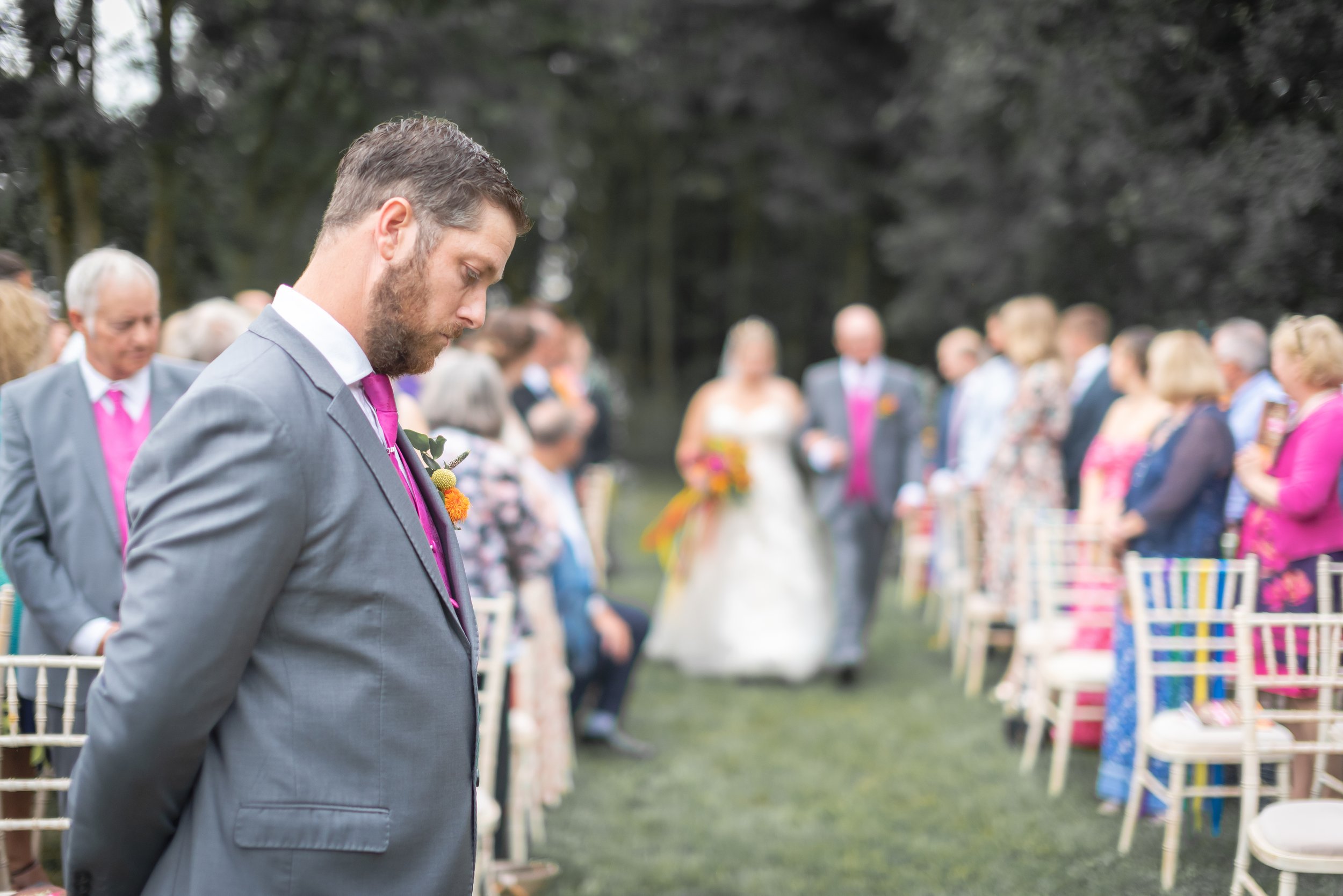 A man in a gray suit and pink tie stands with his head bowed and eyes closed at an outdoor wedding ceremony. In the background, a bride and groom walk down an aisle lined with chairs, with guests standing on either side at Thorney Lakes.
