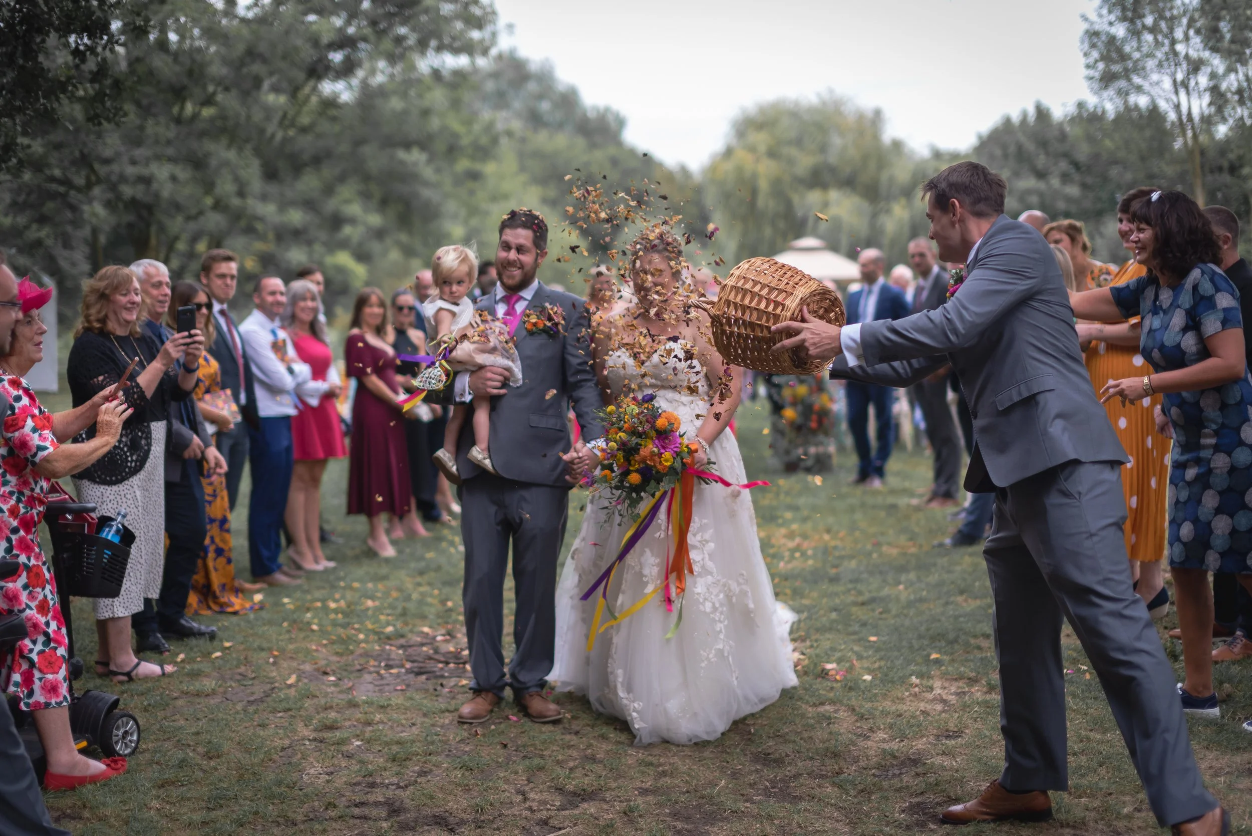 A wedding celebration outdoors where a man in a gray suit is tossing confetti from a basket towards a bride and groom who are holding hands, with a crowd of guests surrounding them at Thorney Lakes.
