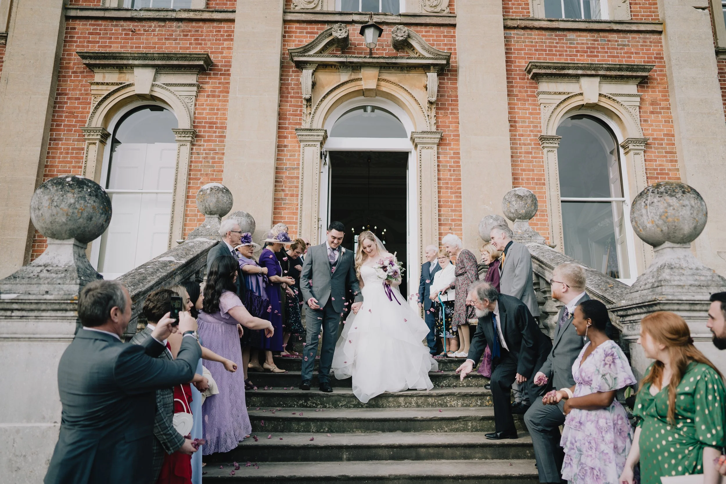 Bride and groom walking through a joyful confetti throw in front of the grand entrance at Crowcombe Court.