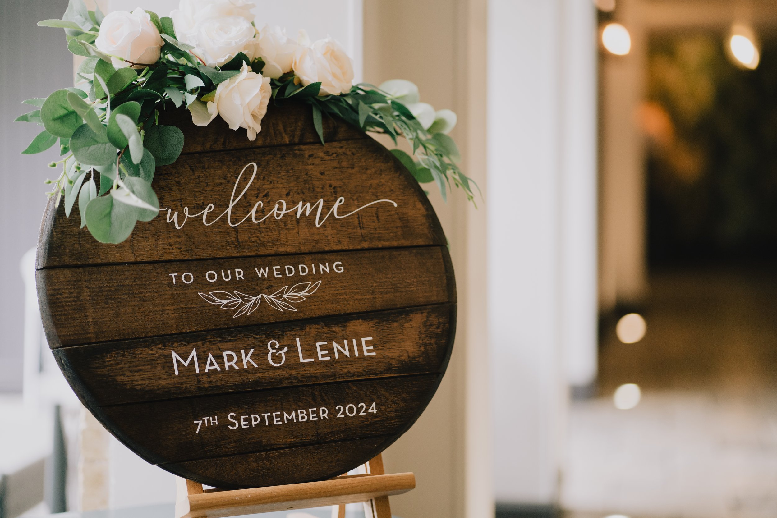 Wooden wedding welcome sign decorated with white flowers and green foliage, reading 'Welcome to our wedding Mark & Lenie 7th September 2024' in white text at Manor House Moreton