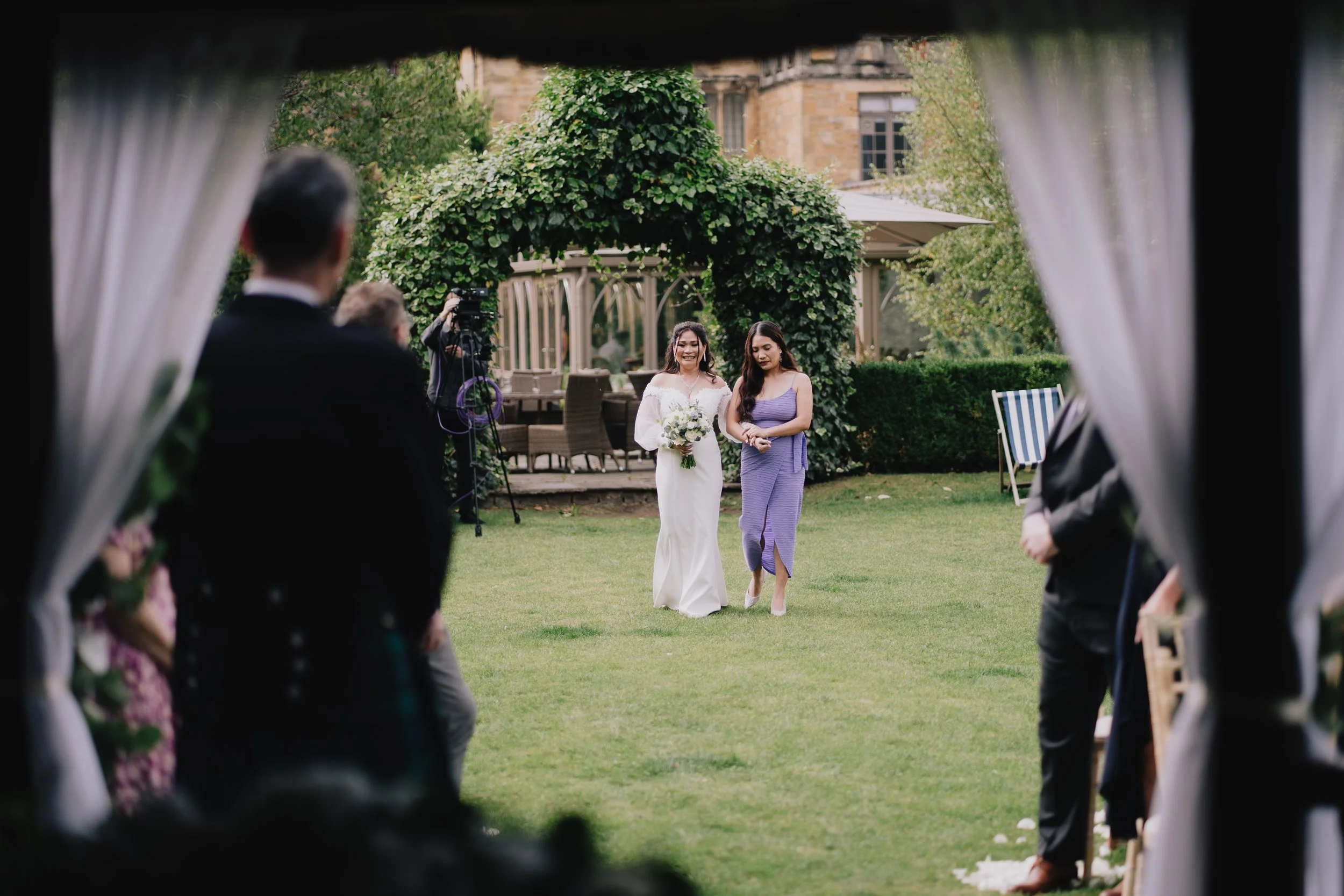 A bride in a white wedding dress and a bridesmaid in a purple gown walking down the aisle during an outdoor wedding ceremony, captured from inside a tent with curtains, with guests and a videographer in the background taken by Cotswold Photographer