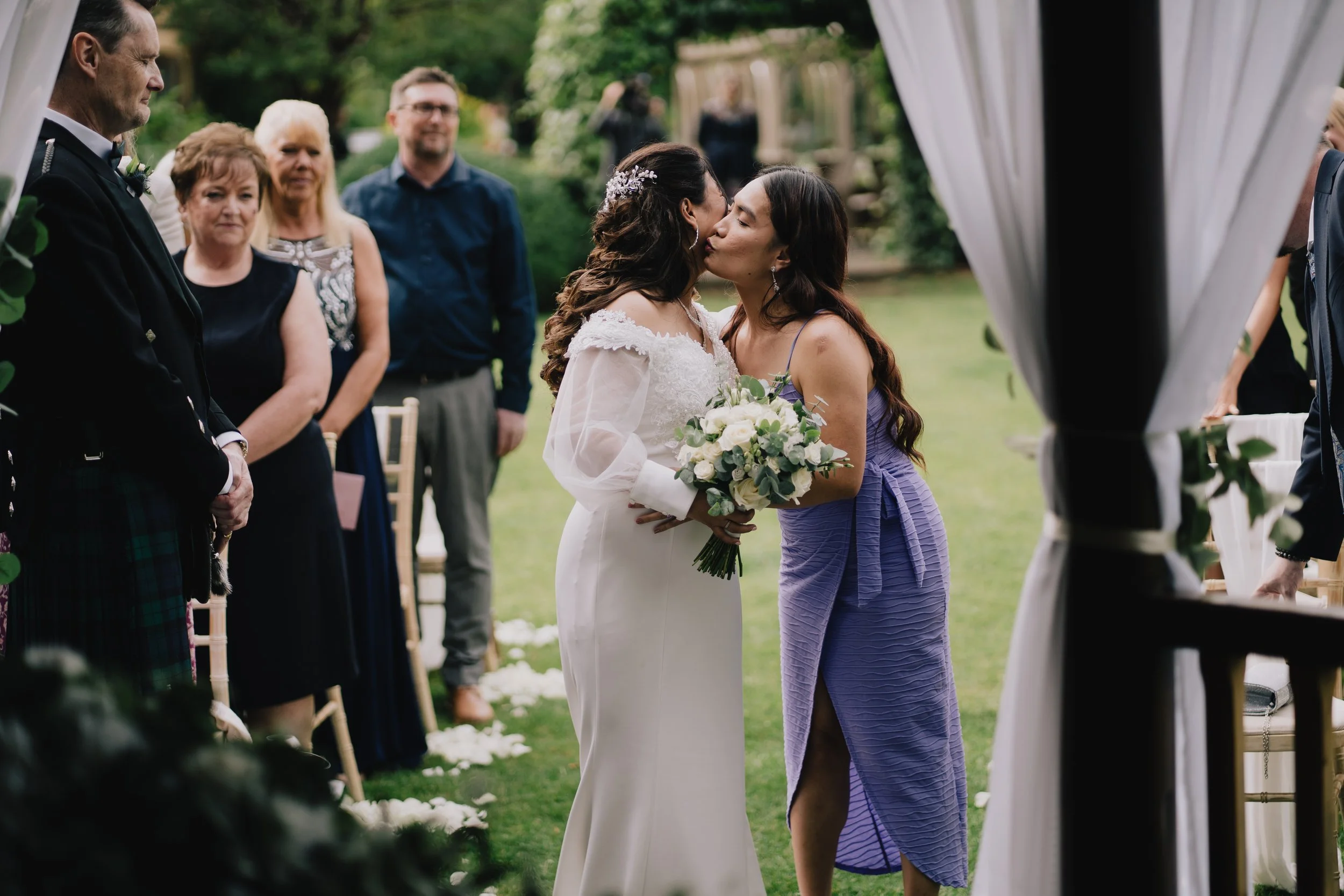 Two women, one in a white wedding dress and the other in a purple dress, share a kiss during a wedding ceremony outdoors, with guests watching taken by Cotswold Wedding Photographer