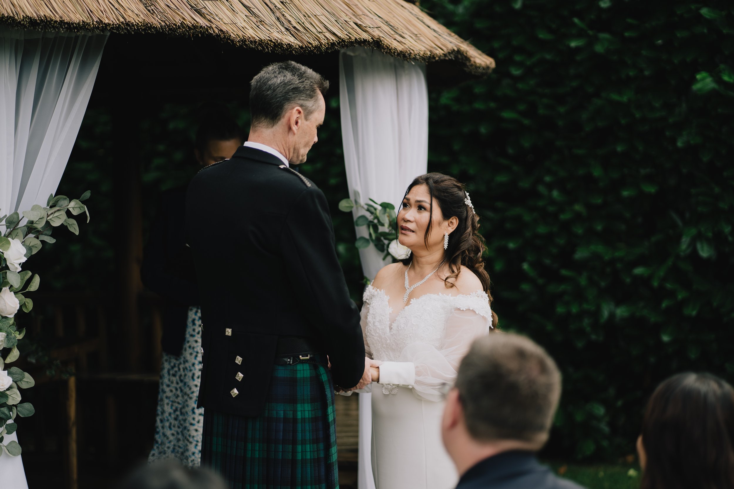 tCotswold Wedding Photographer captures bride and groom holding hands during a wedding ceremony, exchanging vows under a white curtained arbor with greenery in the background.