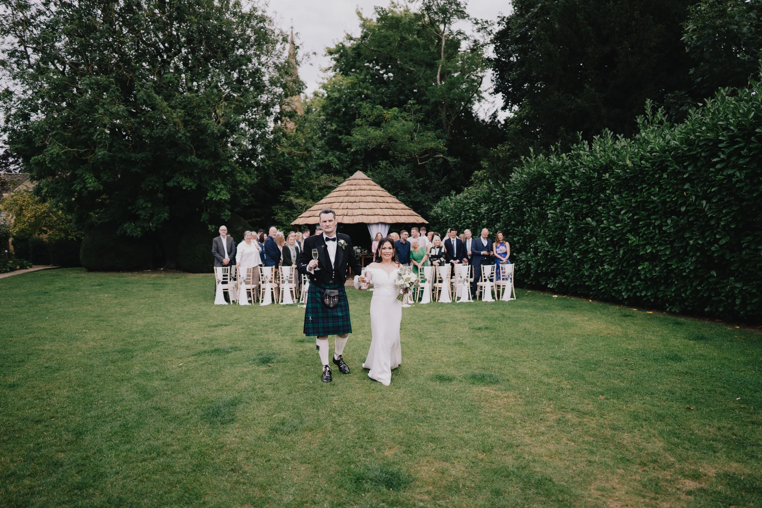 A newlywed couple walking on the grass at their outdoor wedding ceremony, with guests standing behind them in front of a small thatched-roof pavilion and surrounded by trees and bushes at Manor House, Moreton-in-Marsh.