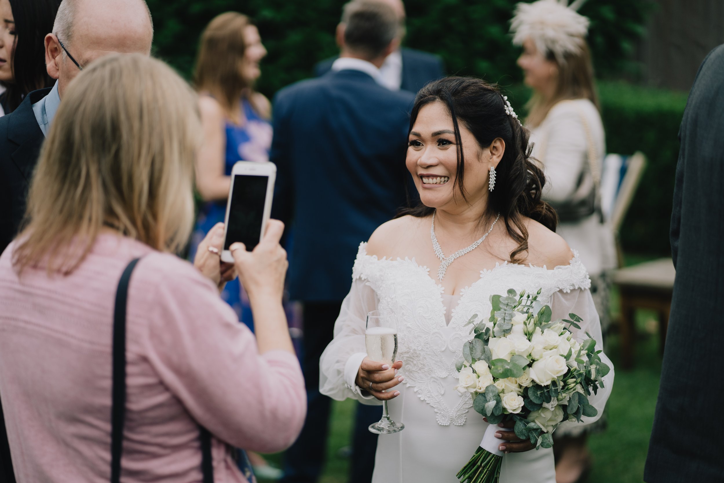 Bride in wedding dress holding a bouquet and glass of Champagne, smiling for a photo taken by another woman, at a wedding reception outdoors at Manor House, Moreton.