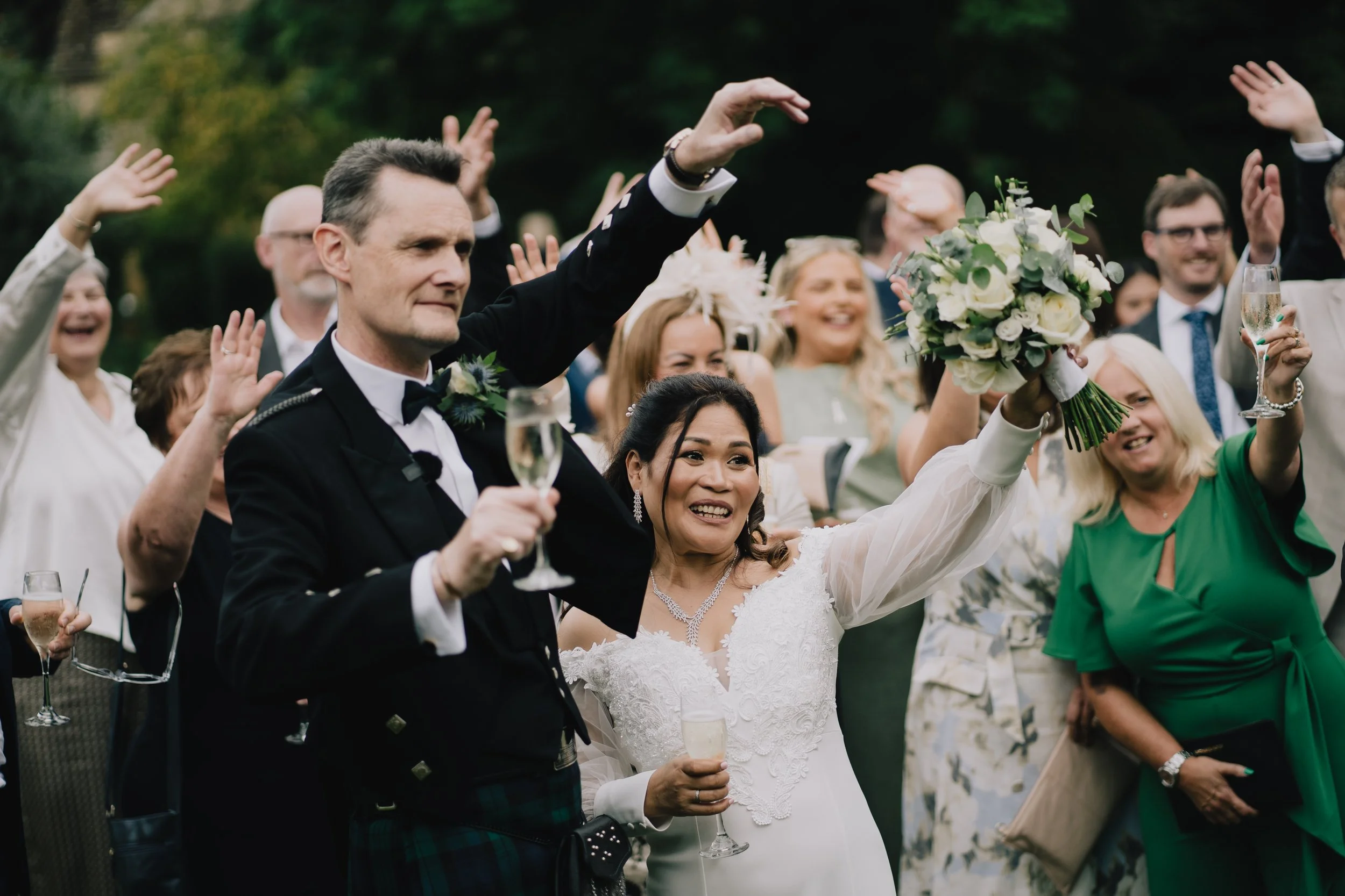 Bride and groom celebrating with guests at a wedding, holding glasses of champagne, outside, with guests smiling, some raising their hands, and the bride holding a large bouquet of white flowers during wedding in Cotswolds.