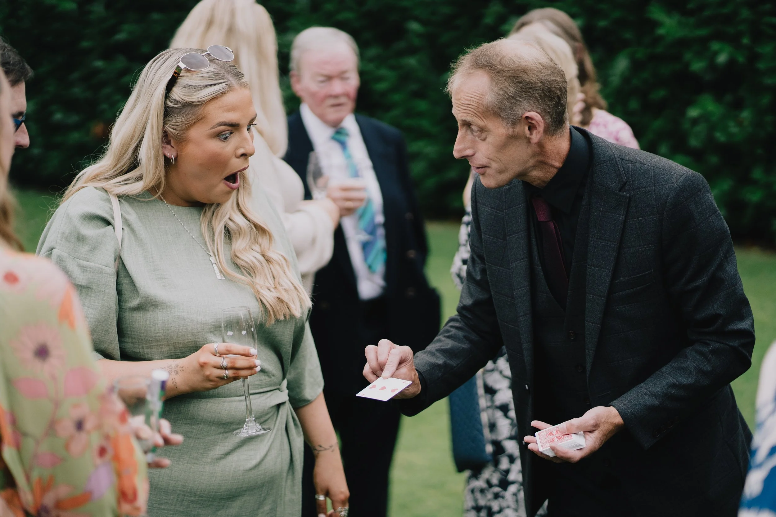 Brett Sirrell, Magician shuffling a deck of playing cards for a guest in a gray dress holding a glass of wine during an outdoor gathering.