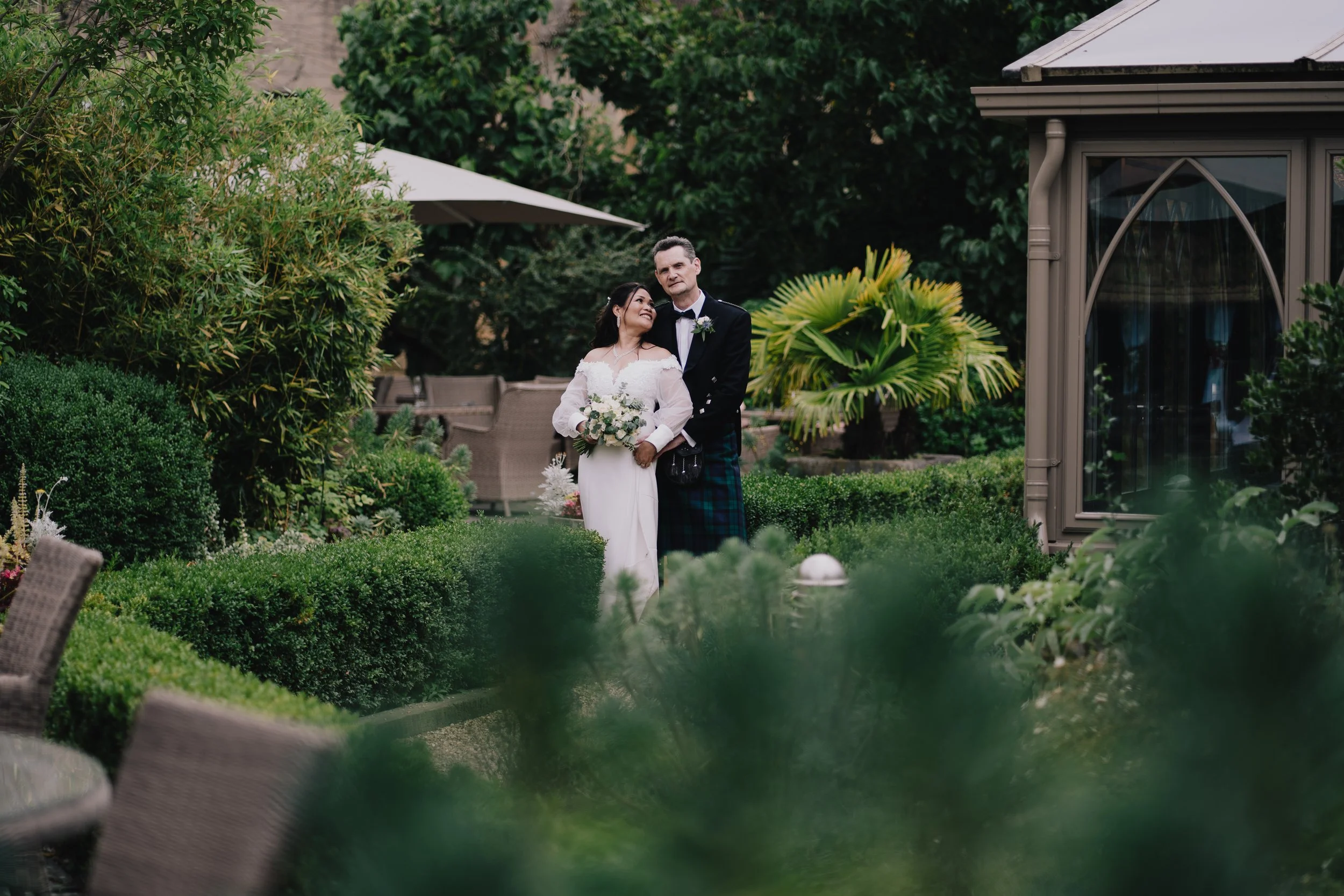 A bride and groom stand together in a lush garden, with the bride holding a bouquet of flowers and smiling at the groom, who is dressed in traditional Scottish attire taken by Cotswold Wedding Photographer.