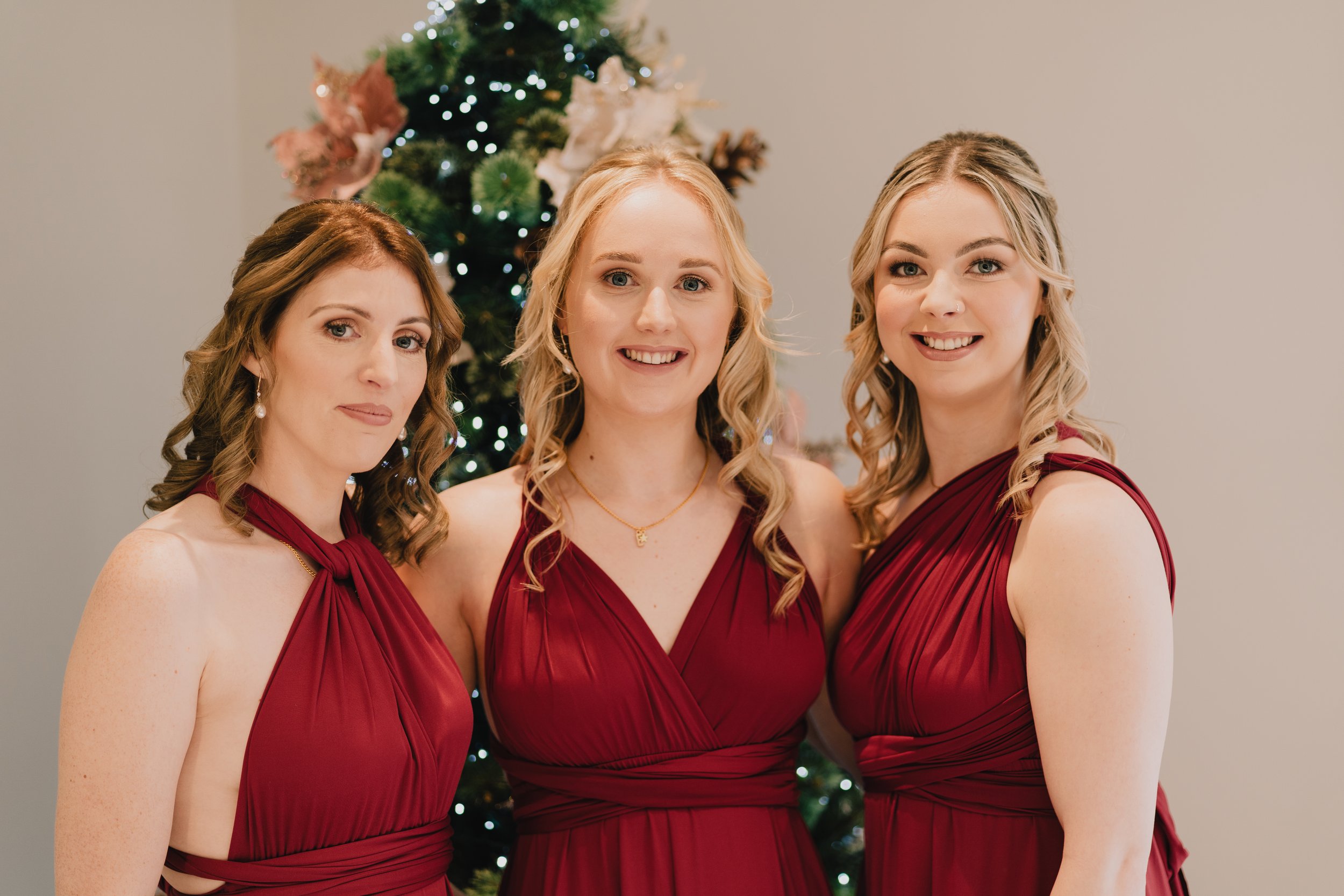 Bridesmaids in matching dresses posing in front of a decorated Christmas tree at Quantock Lakes festive wedding.