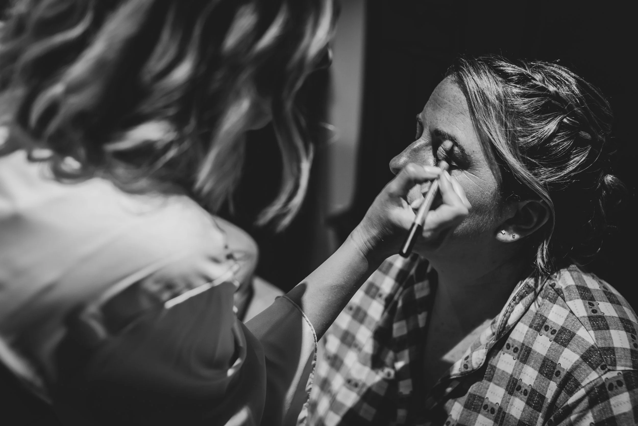 A makeup artist applying eyeshadow to a woman with closed eyes, wearing a checkered shirt, in black and white during prep in Somerset.
