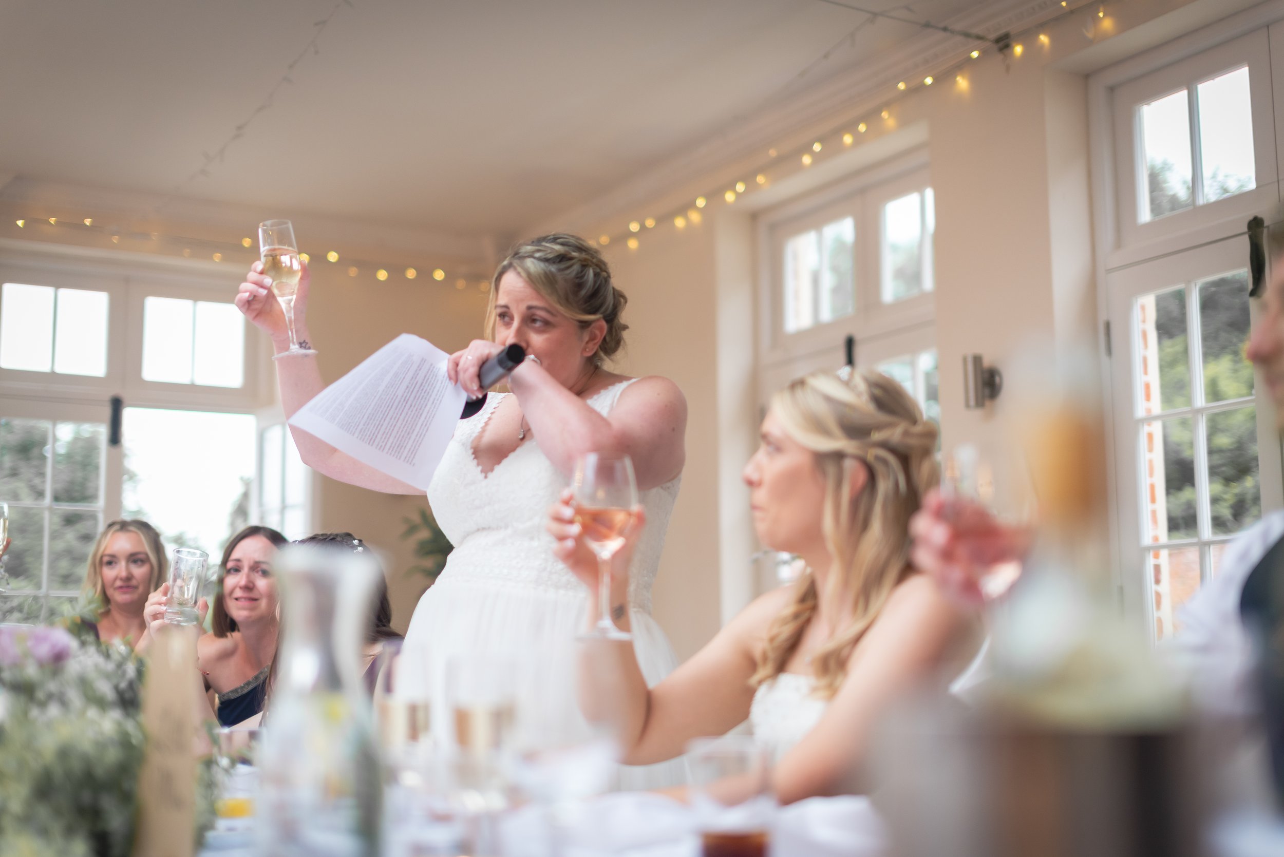A woman in a white dress is giving a speech at a wedding reception, holding a glass of champagne in one hand and a microphone and paper in the other. Several women are seated at the table, holding glasses of pink and clear drinks, and smiling. The room has large windows, string lights, and a festive atmosphere.
