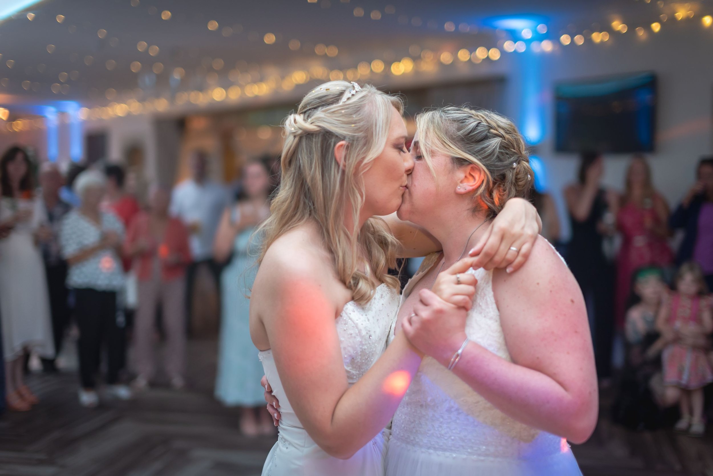 Two women, wearing wedding dresses, sharing a kiss on the dance floor at a during their first dance at Elmhay Park.