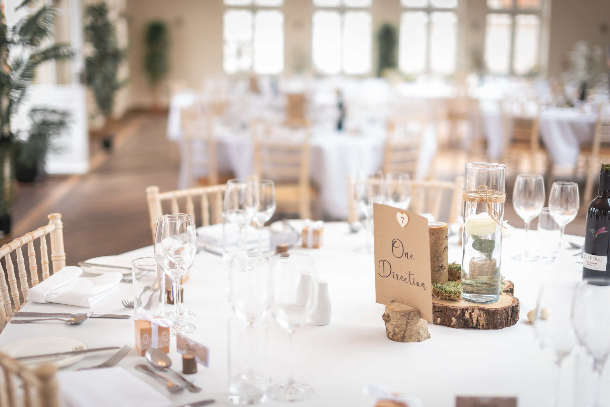 Elegant banquet table set with multiple glasses, white napkins, and cutlery; centerpiece with glass of water, small wooden sign indicating 'One Direction,' and candle in glass holder at Elmhay Park.