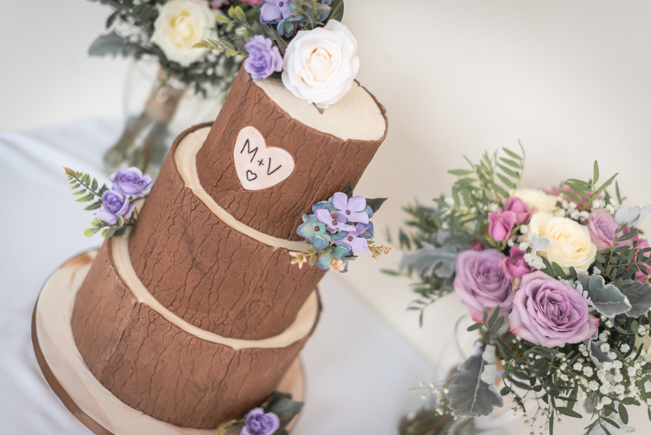 A three-tiered cake designed to look like tree logs, decorated with purple and white flowers, and a heart-shaped sign with the words 'M + V' and a small heart, surrounded by floral arrangements with pink, purple, white roses, and greenery.