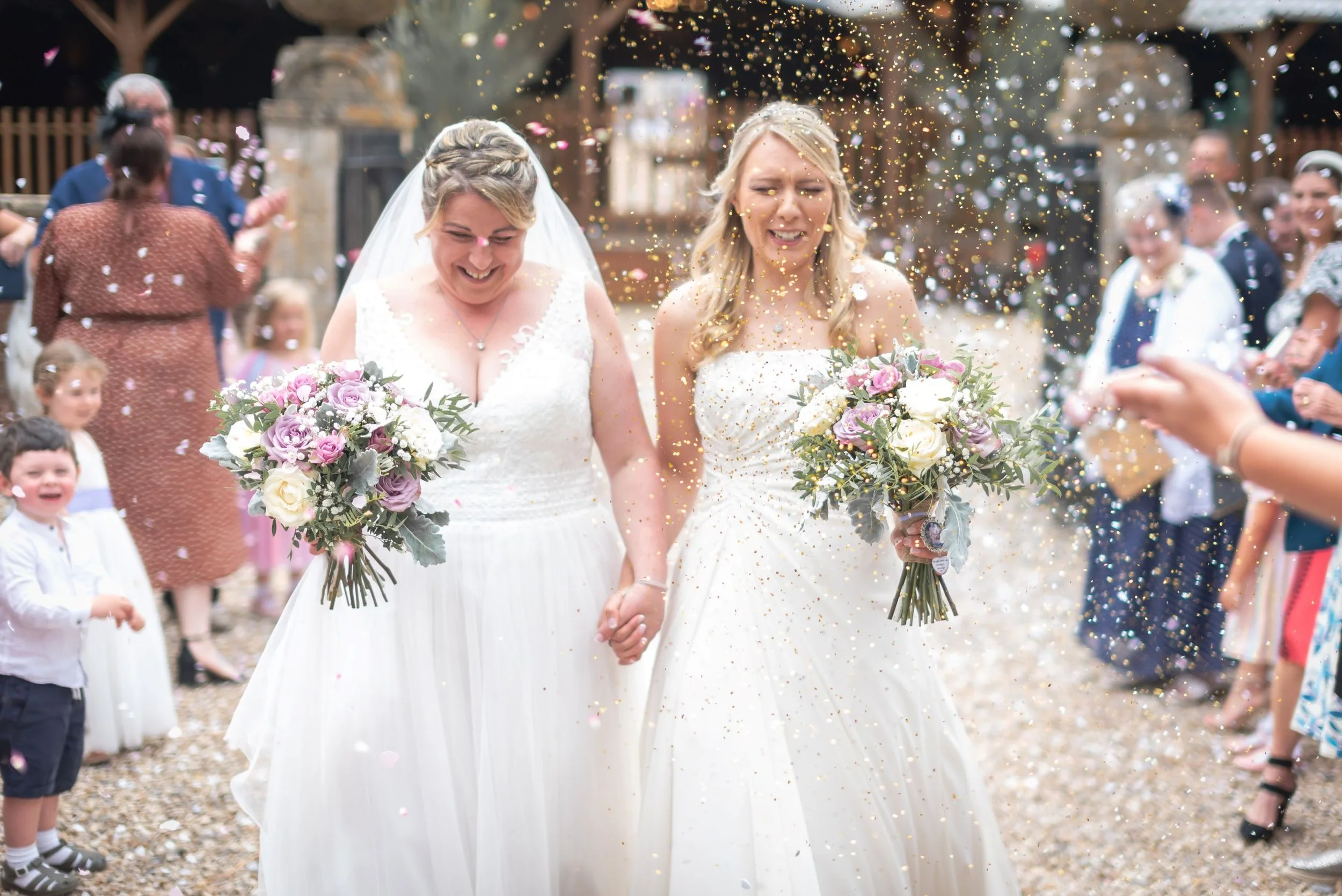 Two brides in white wedding dresses holding hands, walking outdoors with confetti falling around them, surrounded by guests and children celebrating their wedding taken by Bristol wedding photographer.