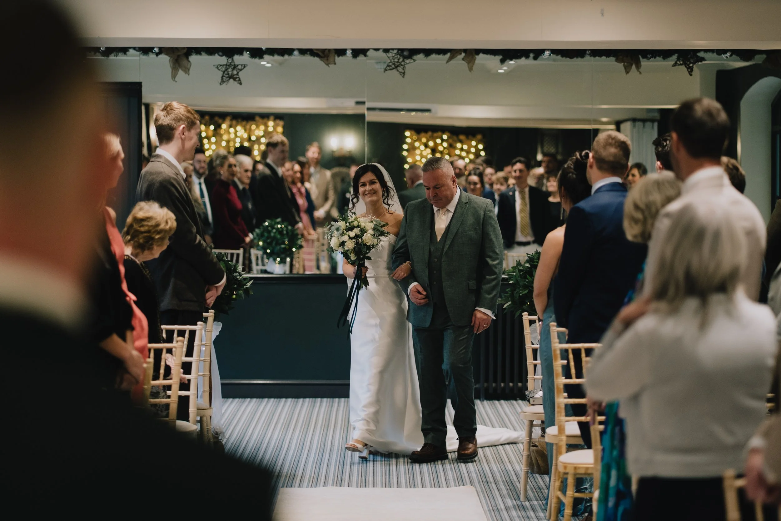 A bride in a white wedding dress holding a bouquet and walking down the aisle with her father at a wedding ceremony, surrounded by seated guests and decorated with Christmas lights at Manor House, Moreton-in-Marsh.