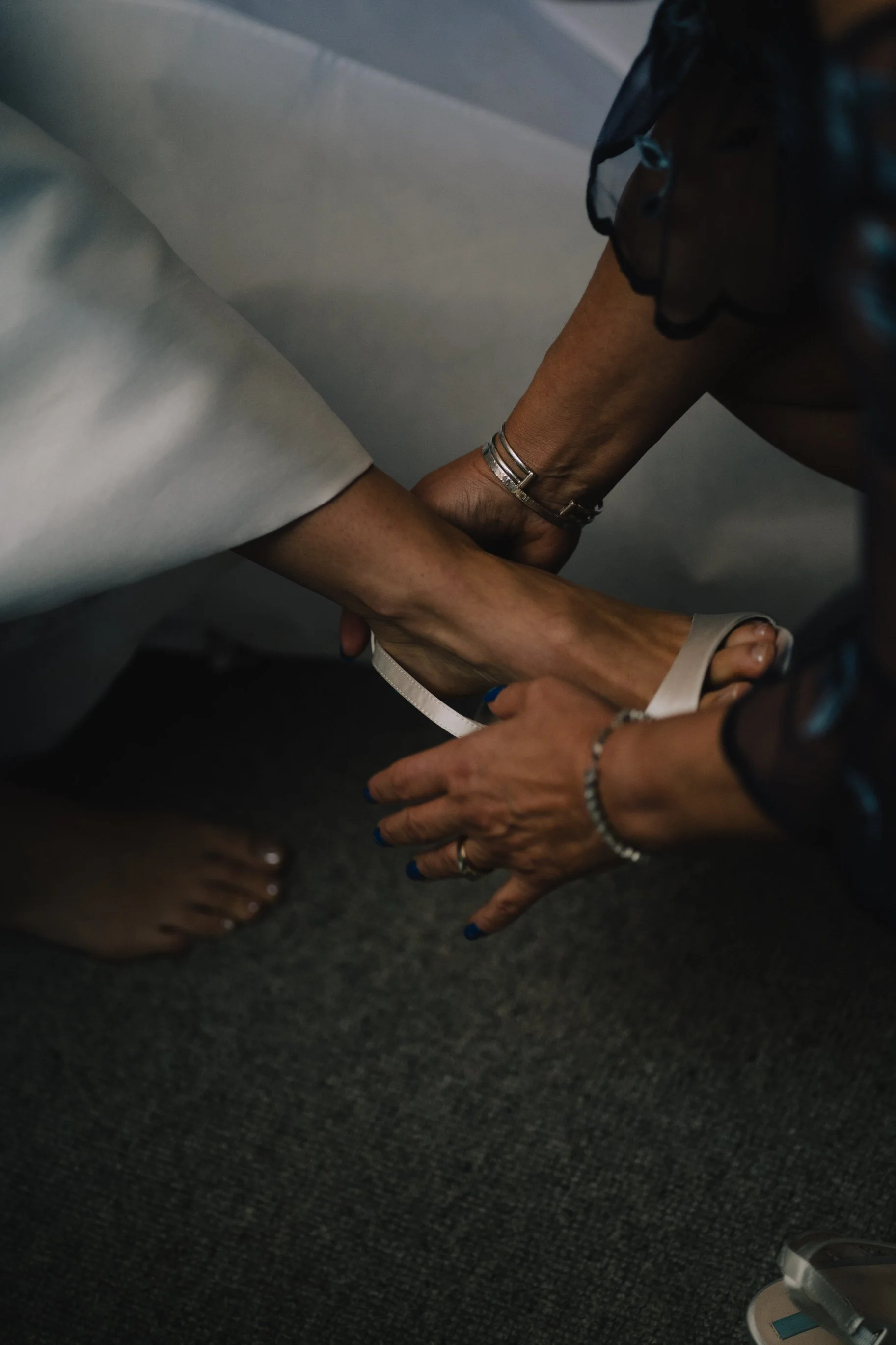 Bride in a white dress being assisted with a white shoe from a woman wearing dark clothing, jewelry, and a bracelet, on a gray carpeted floor.
