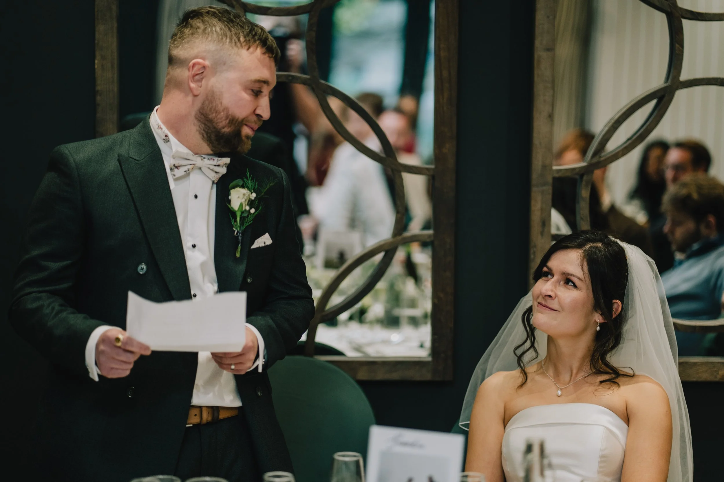 Groom giving a speech at a wedding reception, standing next to the bride who is sitting and listening with a smile in Cotswolds.