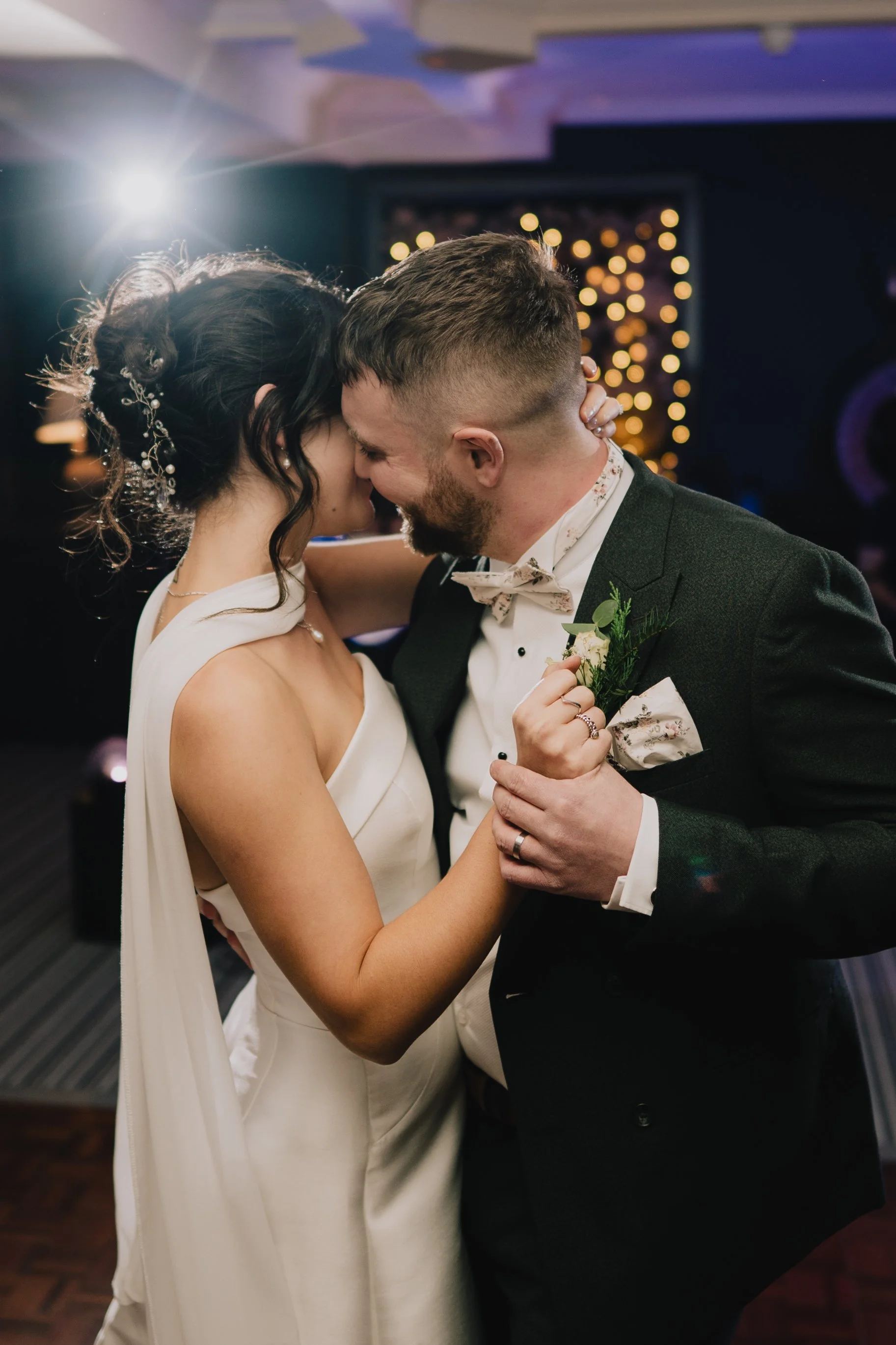 A newlywed couple sharing their first dance at their wedding reception at Manor House in Moreton-in-Marsh, dressed in wedding attire, with warm lighting and blurred decorative lights in the background.