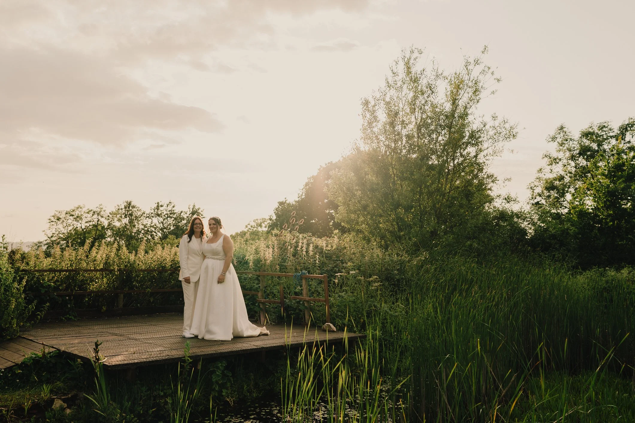 Brides embracing by the pond at Folly Farm during sunset golden hour