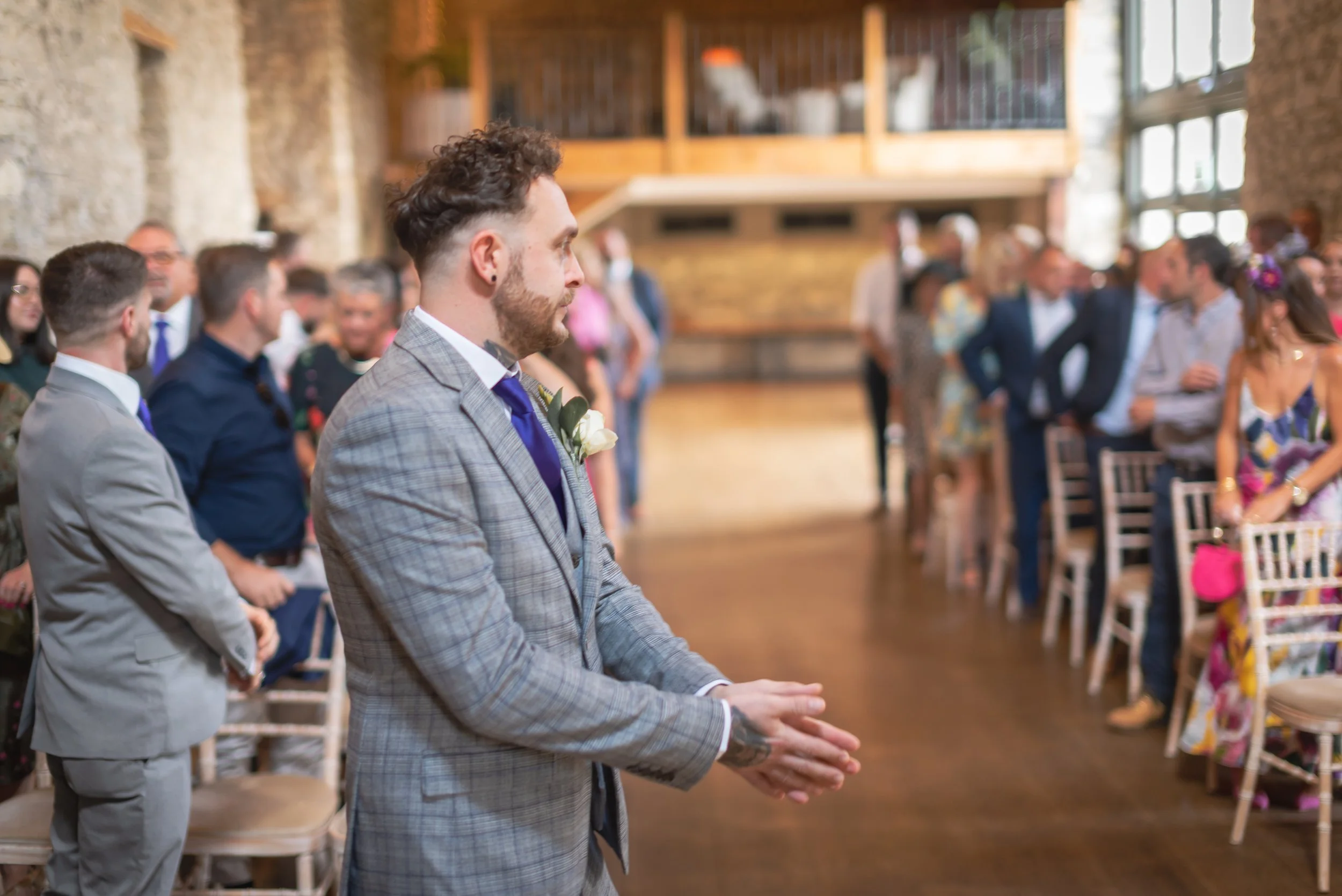 Groom in a gray plaid suit standing with hands clasped at a wedding ceremony, surrounded by guests in a rustic venue with exposed brick and wooden accents.