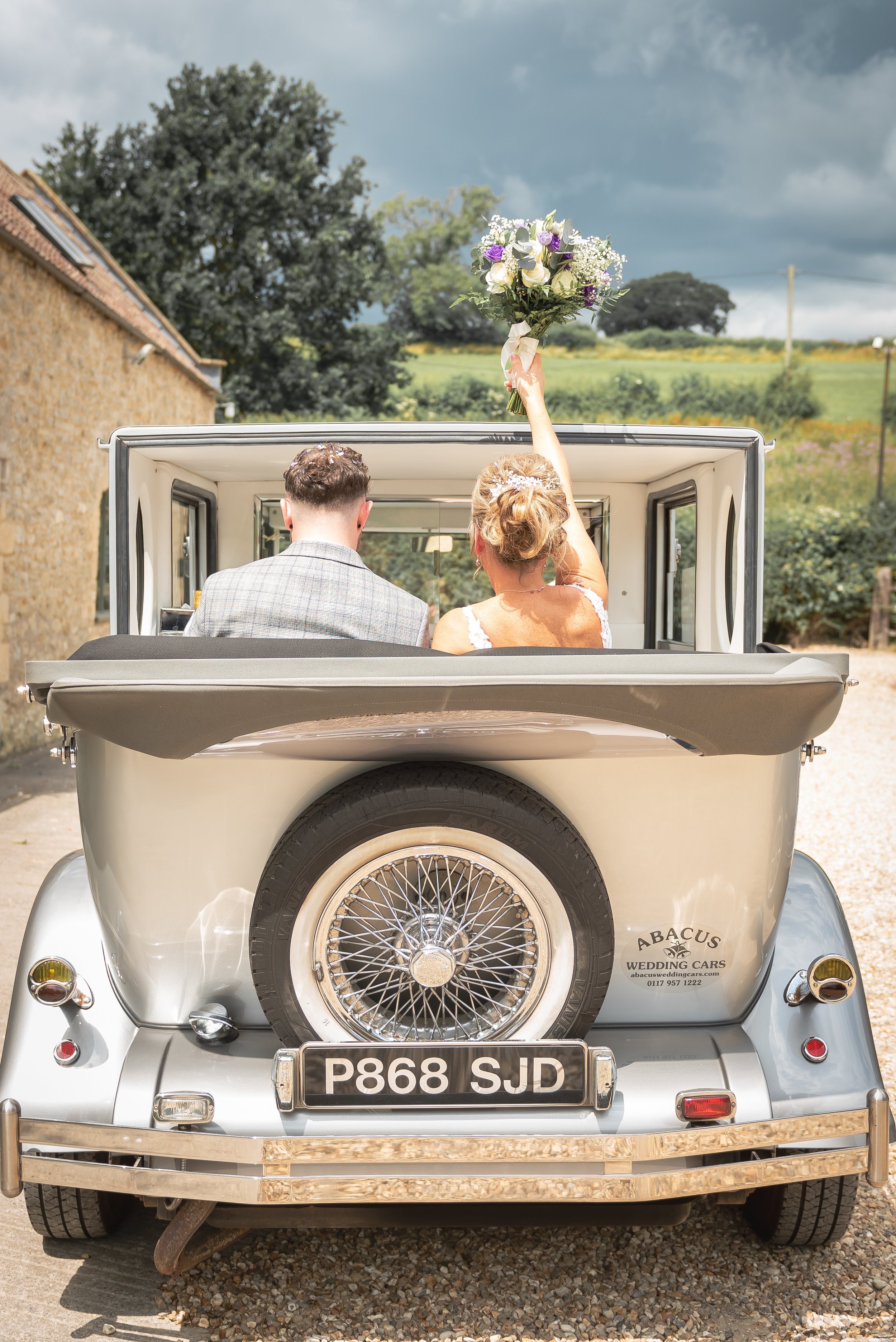 Bride and groom sitting in the back of a vintage car, with the bride holding a bouquet and raising it in the air, outdoors with a countryside backdrop after leaving Priston Mill.