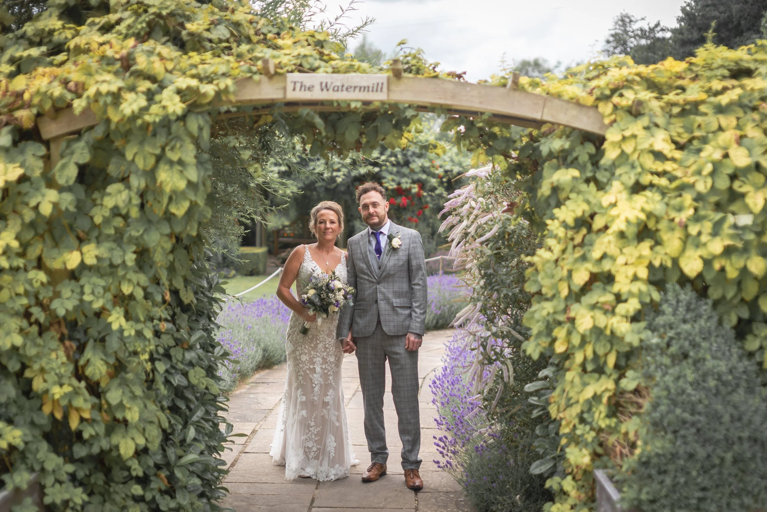 A bride and groom standing hand in hand under an archway labeled 'The Watermill' in a garden with purple flowers and greenery, celebrating their wedding at Priston Mill.