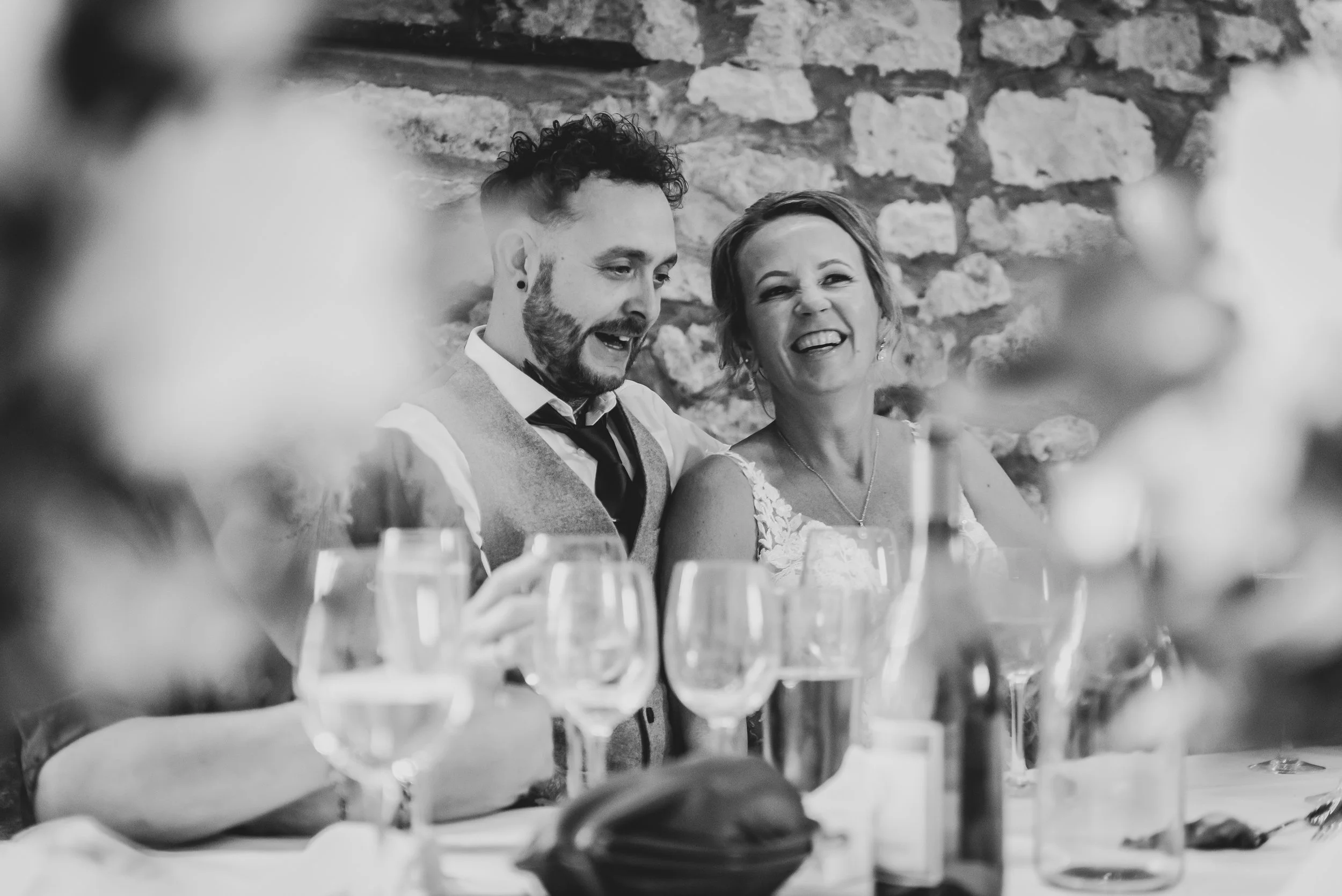 Black and white photo of a bride and groom sitting at a wedding banquet table, smiling and laughing. The background features a brick wall, and there are wine glasses and a flower on the table in front of them.