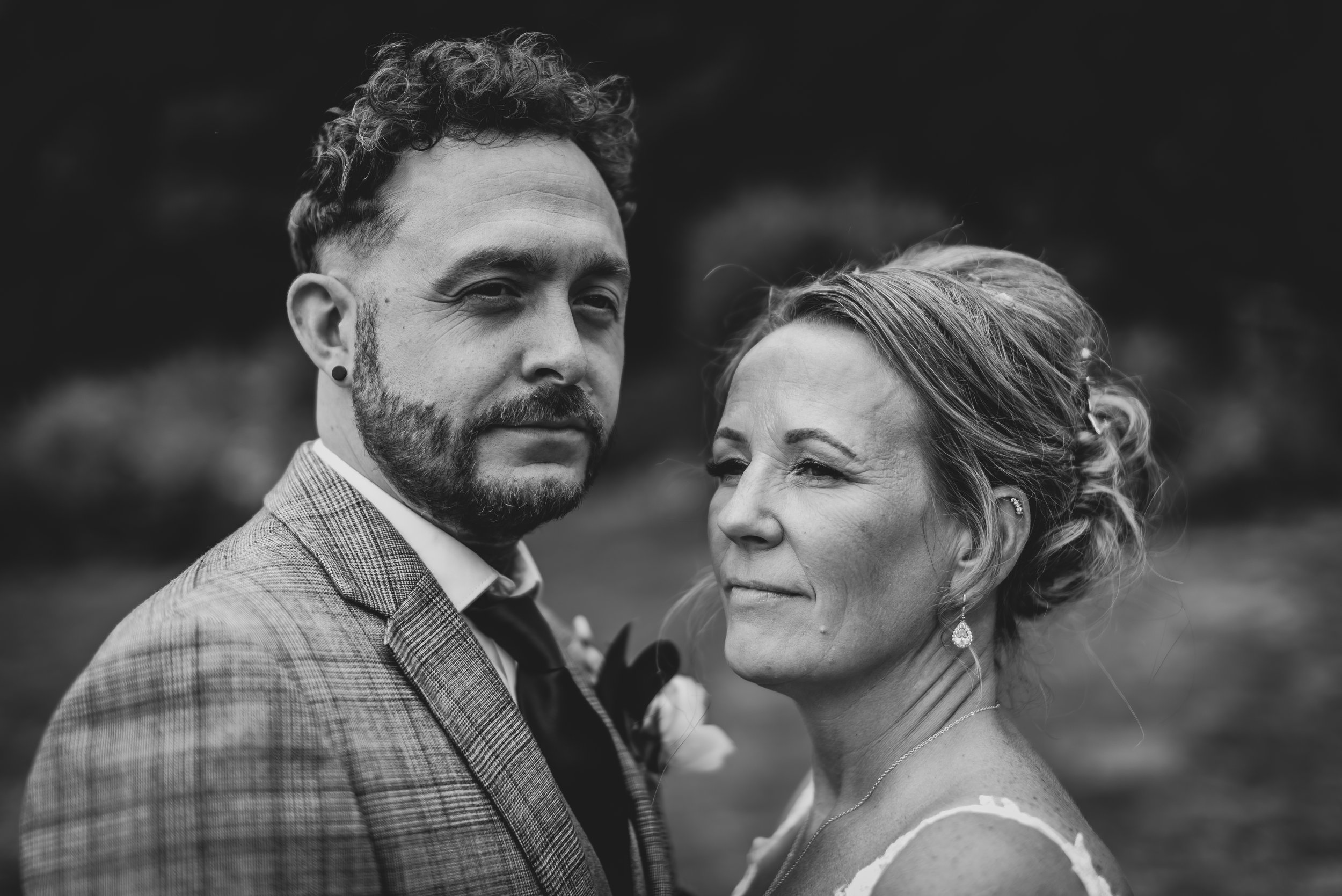 A black and white photograph of a couple, likely on their wedding day, standing outdoors with a blurred natural background. The man has curly hair, a beard, and wears a checked suit with a boutonniere. The woman has her hair styled in an updo, wears earrings, a necklace, and a dress with thin straps.