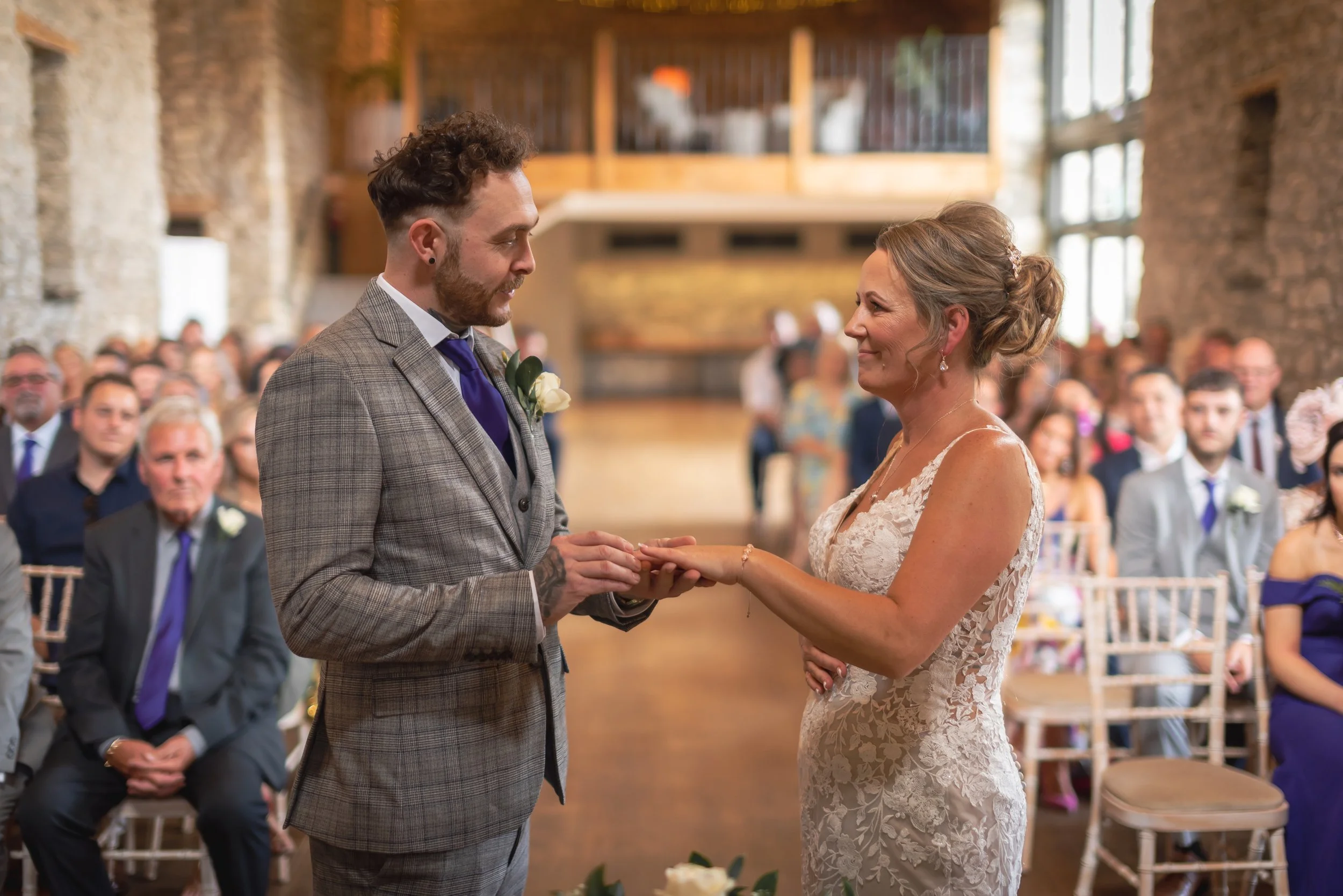 A bride and groom exchanging rings during a wedding ceremony in a rustic indoor venue filled with seated guests captured by Priston Mill Wedding Photographer
