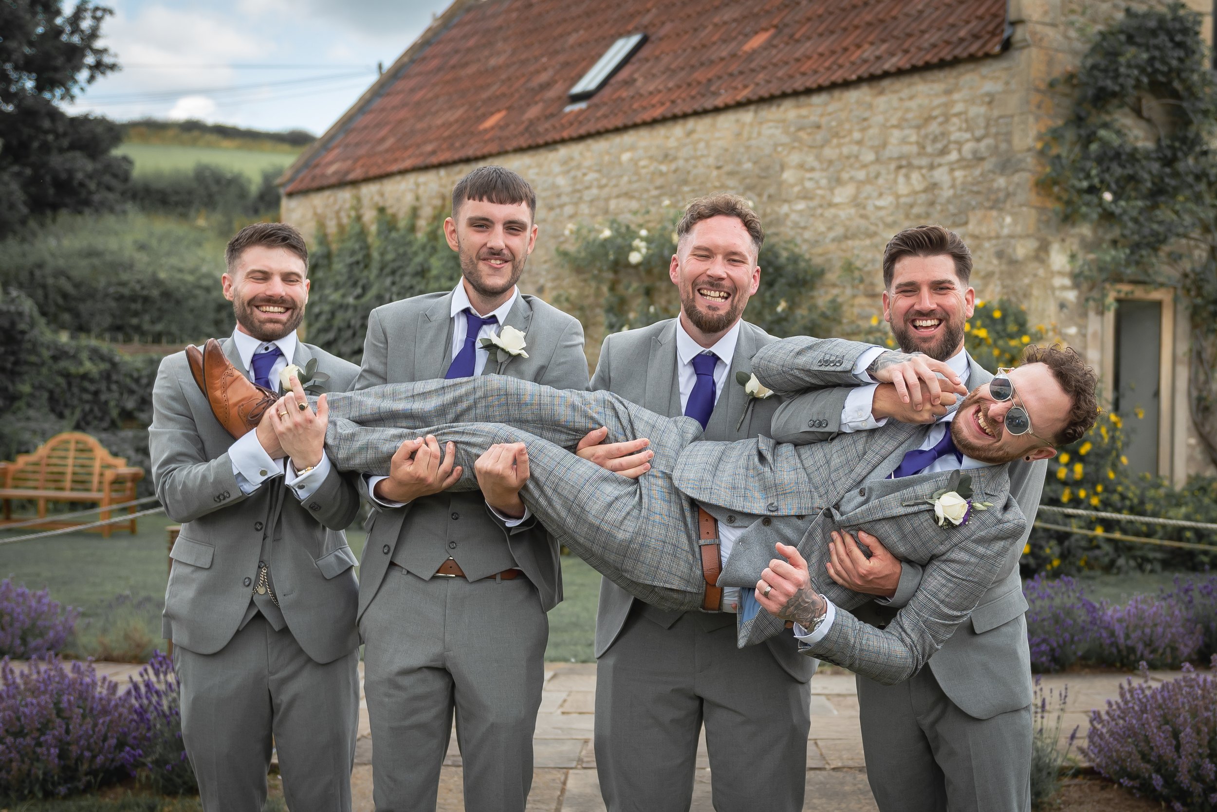 Group of five men in gray suits holding the groom in a plaid suit horizontally, smiling at a wedding by Priston Mill Photographer