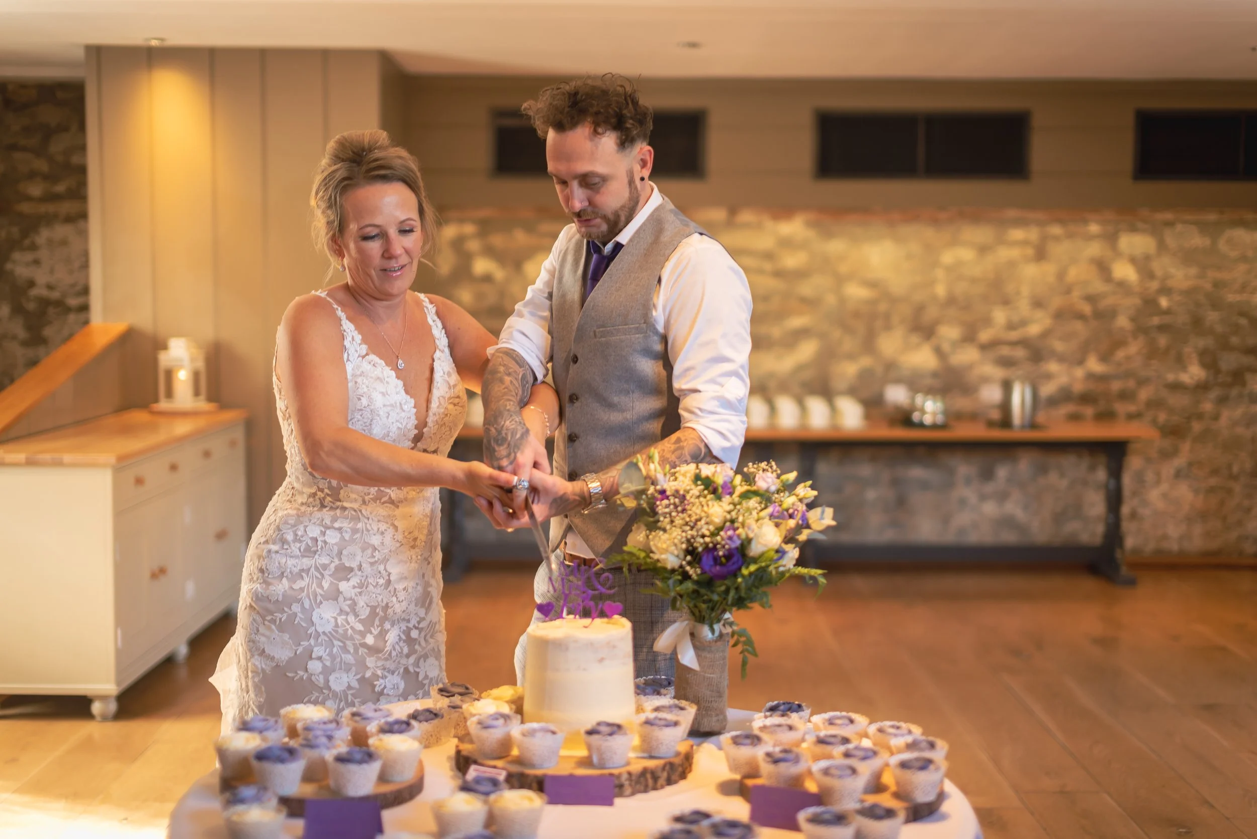 A bride and groom cutting a wedding cake together at their reception, with cupcakes on the table and a floral arrangement nearby during the evening reception at Priston Mill