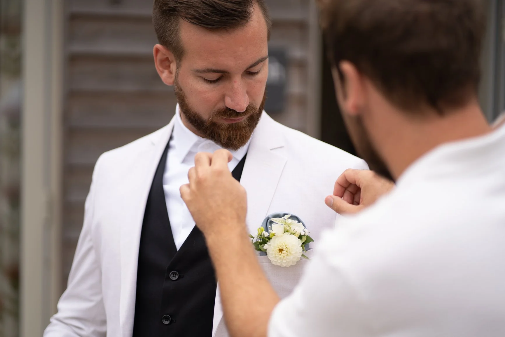A groom in a white tuxedo with a flower boutonniere is being pinned by best man at Oxleaze Barn.