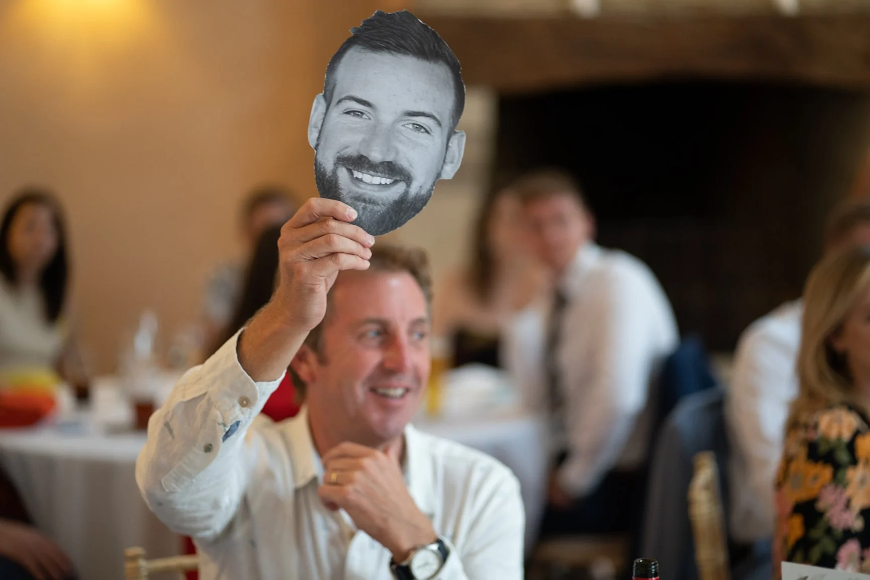 Man smiling and holding a large cutout of a grooms face at a social gathering or party at Oxleaze Barn.