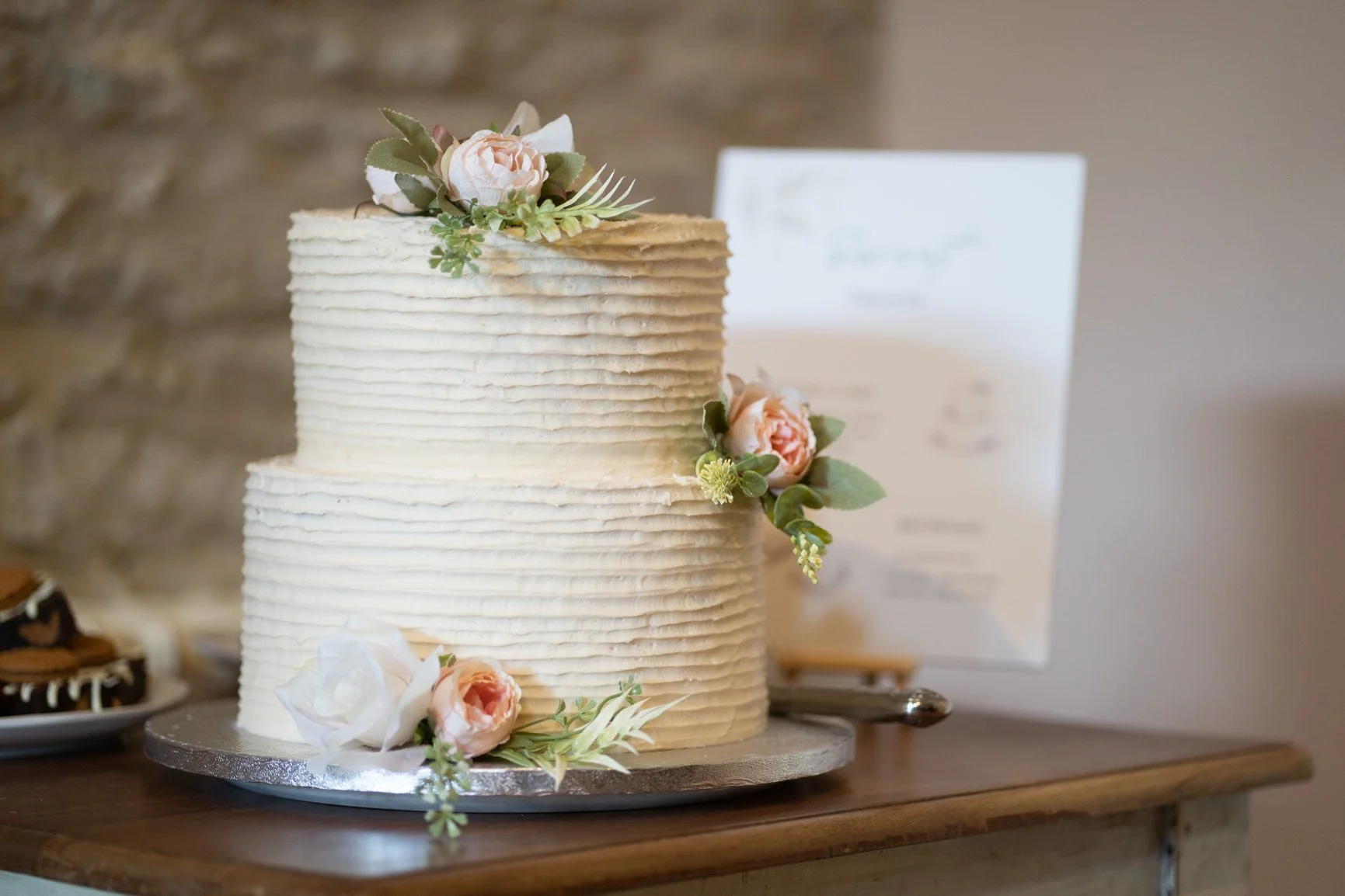 A two-tiered white wedding cake made by Rosie Bear Cake decorated with pink and white flowers and green foliage, placed on a silver cake stand on a wooden table at Oxleaze Barn.