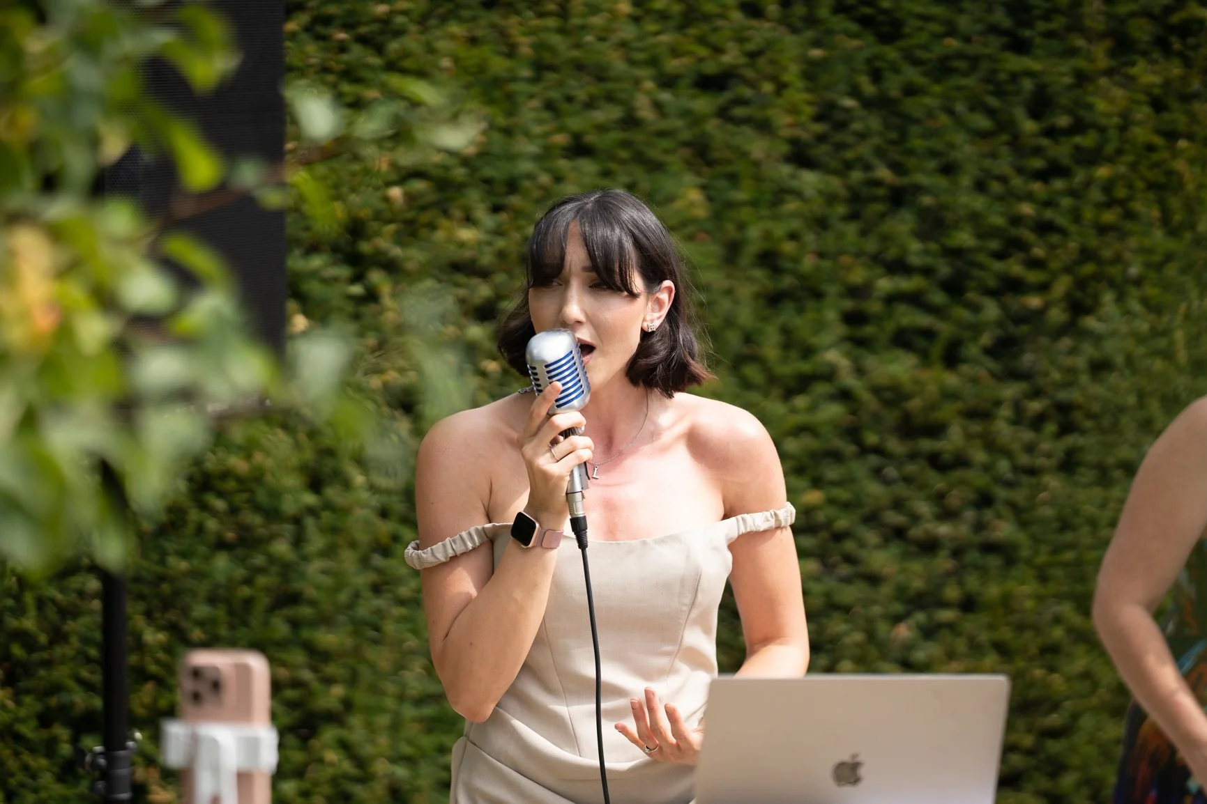 Lisa Mioni in a beige off-shoulder dress singing into a vintage microphone outdoors with a laptop and greenery in the background  at Oxleaze Barn.