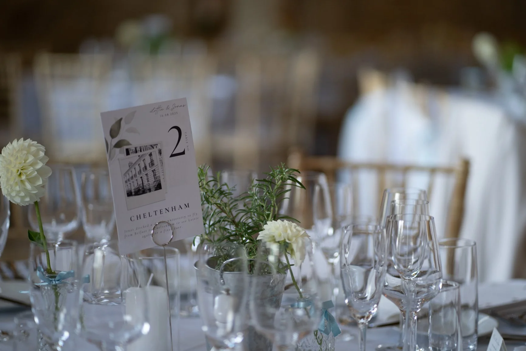 Elegant table setting with white roses, green foliage, and multiple clear wine glasses, with a table number card displaying the number 2 and the word 'Cheltenham'  at Oxleaze Barn.