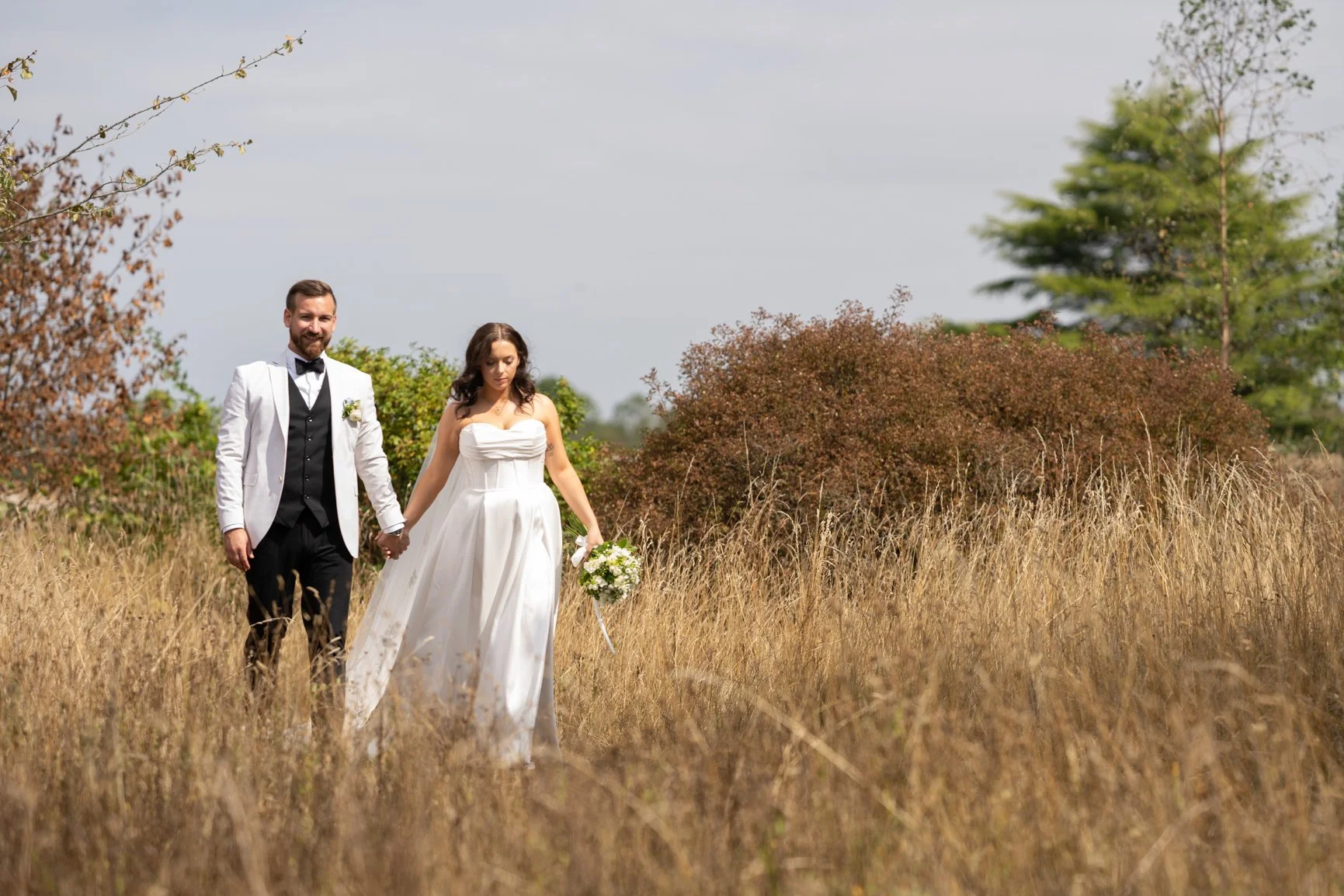 A bride and groom walking hand-in-hand through a field of tall grass, with trees and shrubs in the background  at Oxleaze Barn.