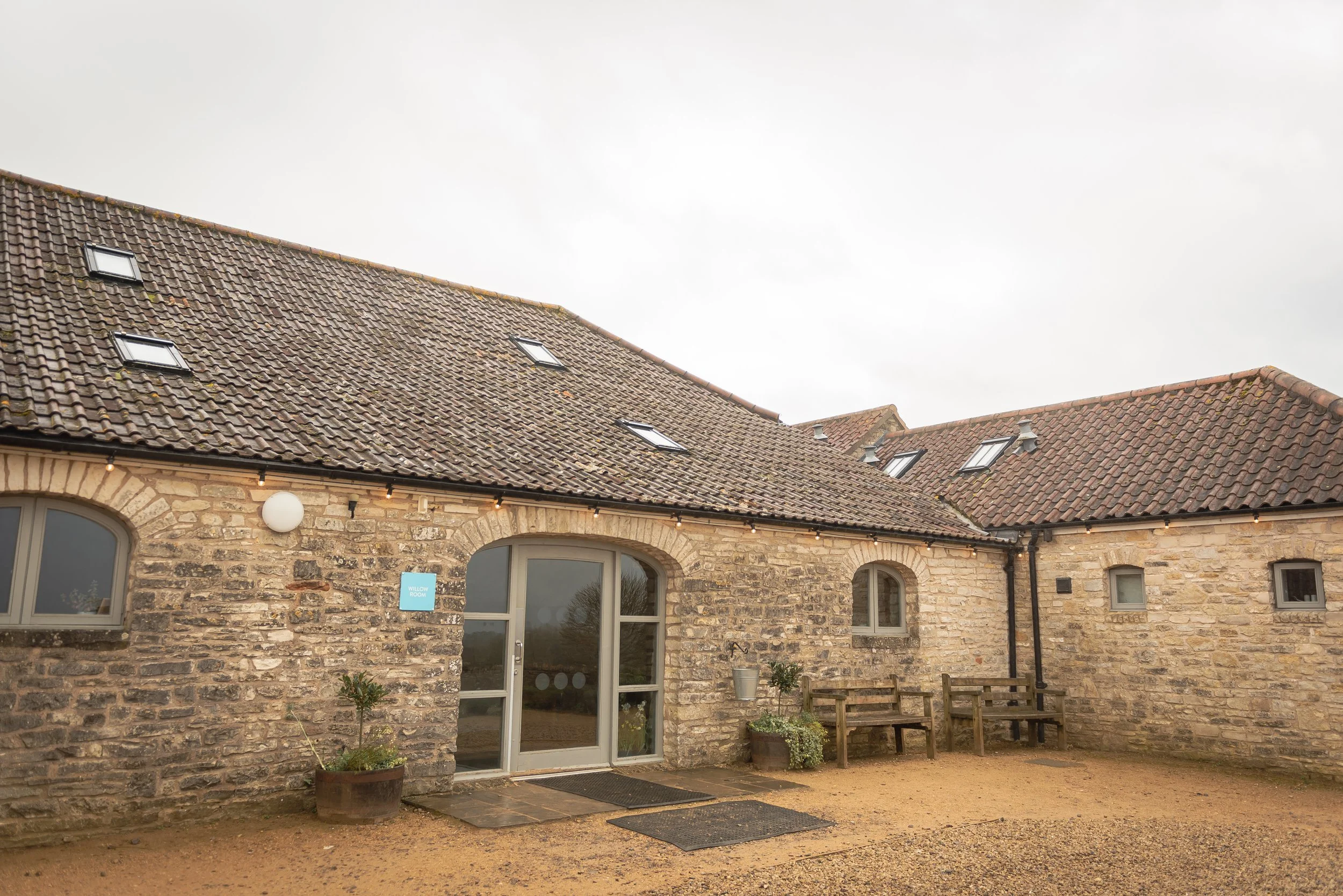 Folly Farm - A rustic stone building with a tiled roof and multiple skylights, featuring a glass door entrance and outdoor seating, under an overcast sky.