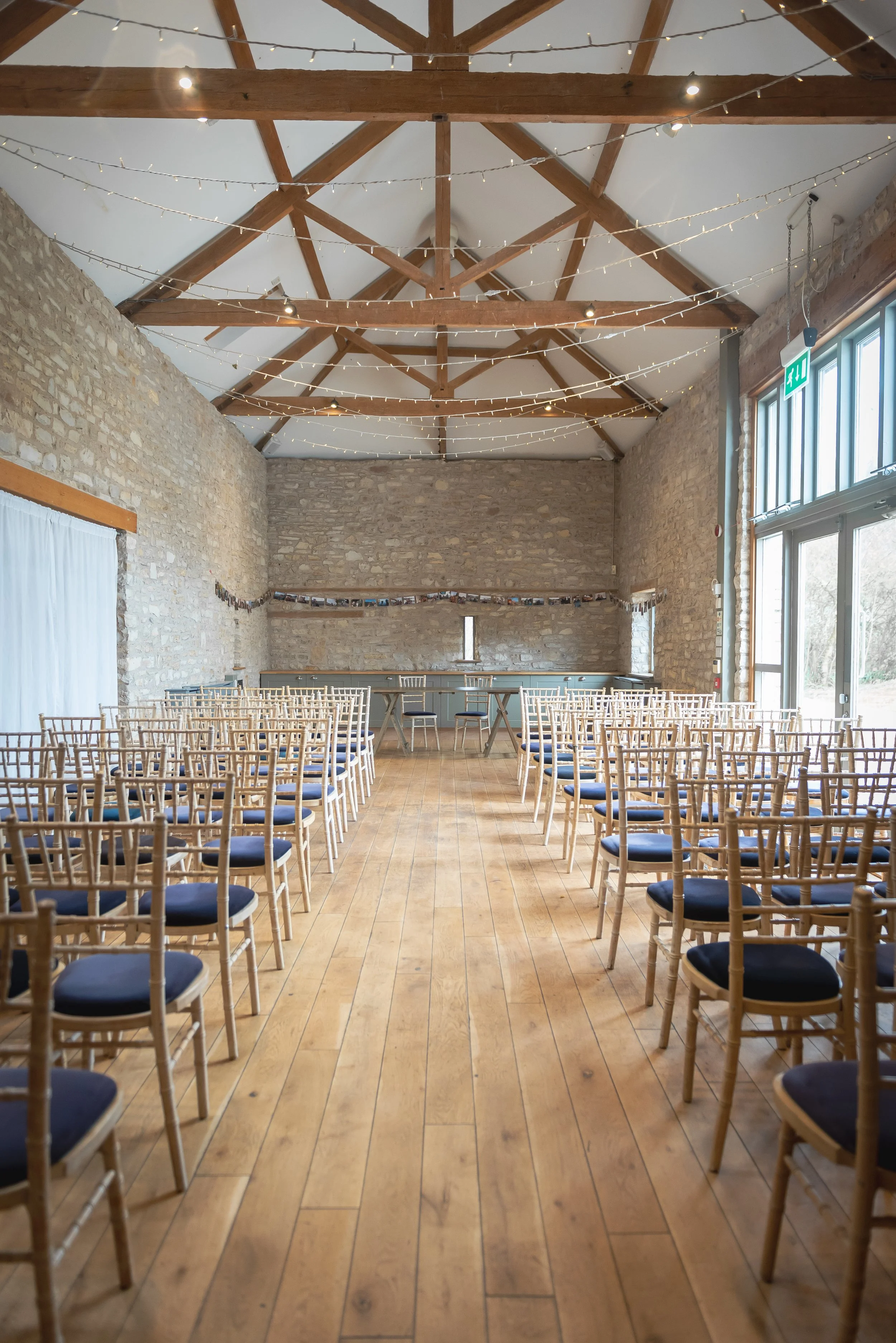 Folly Farm in Bishop Sutton with rows of wooden chairs with dark cushions, arranged facing a raised stage area with tables. The room has high vaulted ceilings with wooden beams and string lights, stone walls, large windows, and a wooden floor.